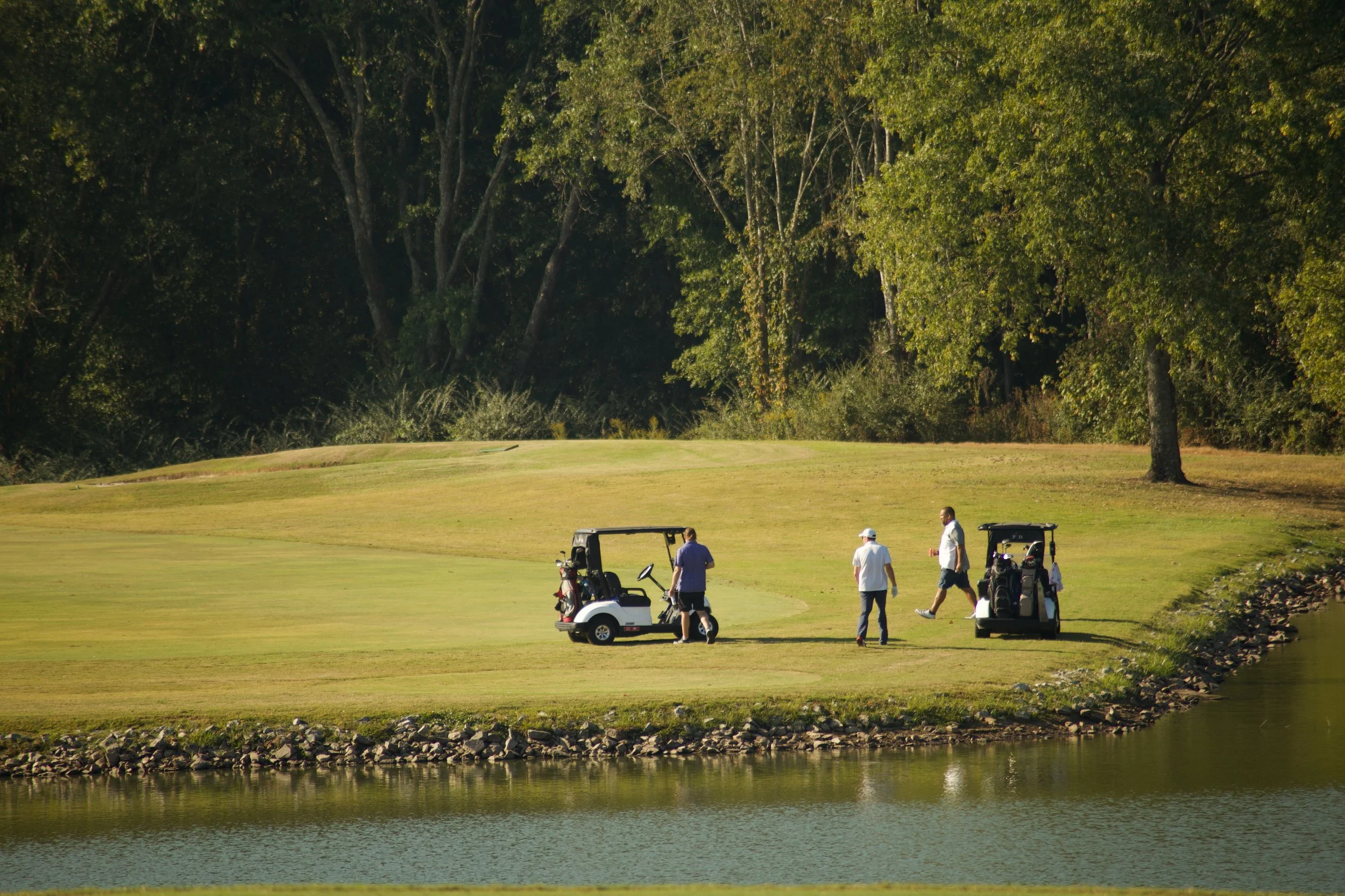 Golf course with four men near golf carts, trees in background, and water in foreground.