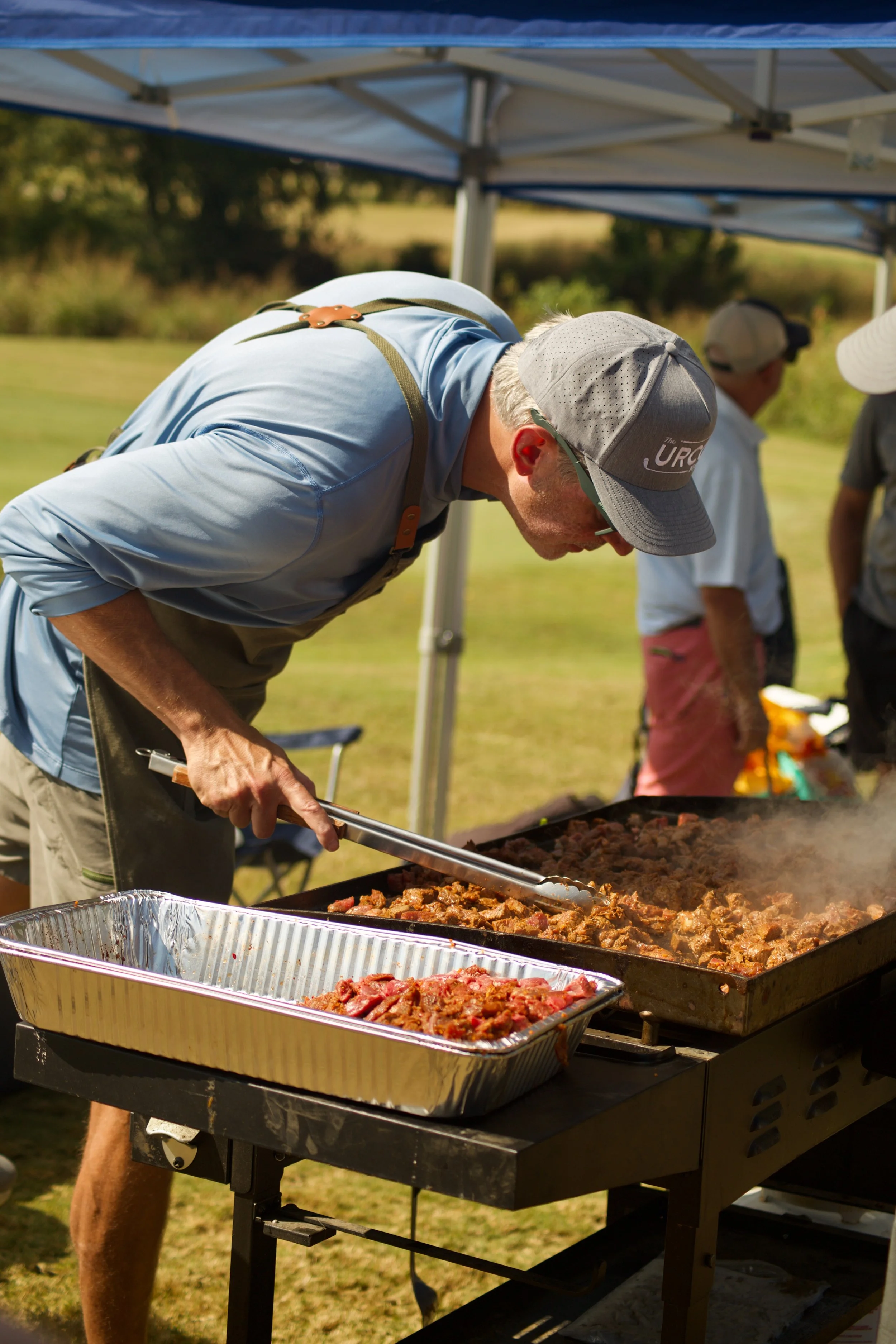 A man wearing a gray cap and sunglasses is serving cooked meat from a large outdoor grill at an outdoor gathering. There are other people nearby, and a canopy tent overhead in a grassy area.
