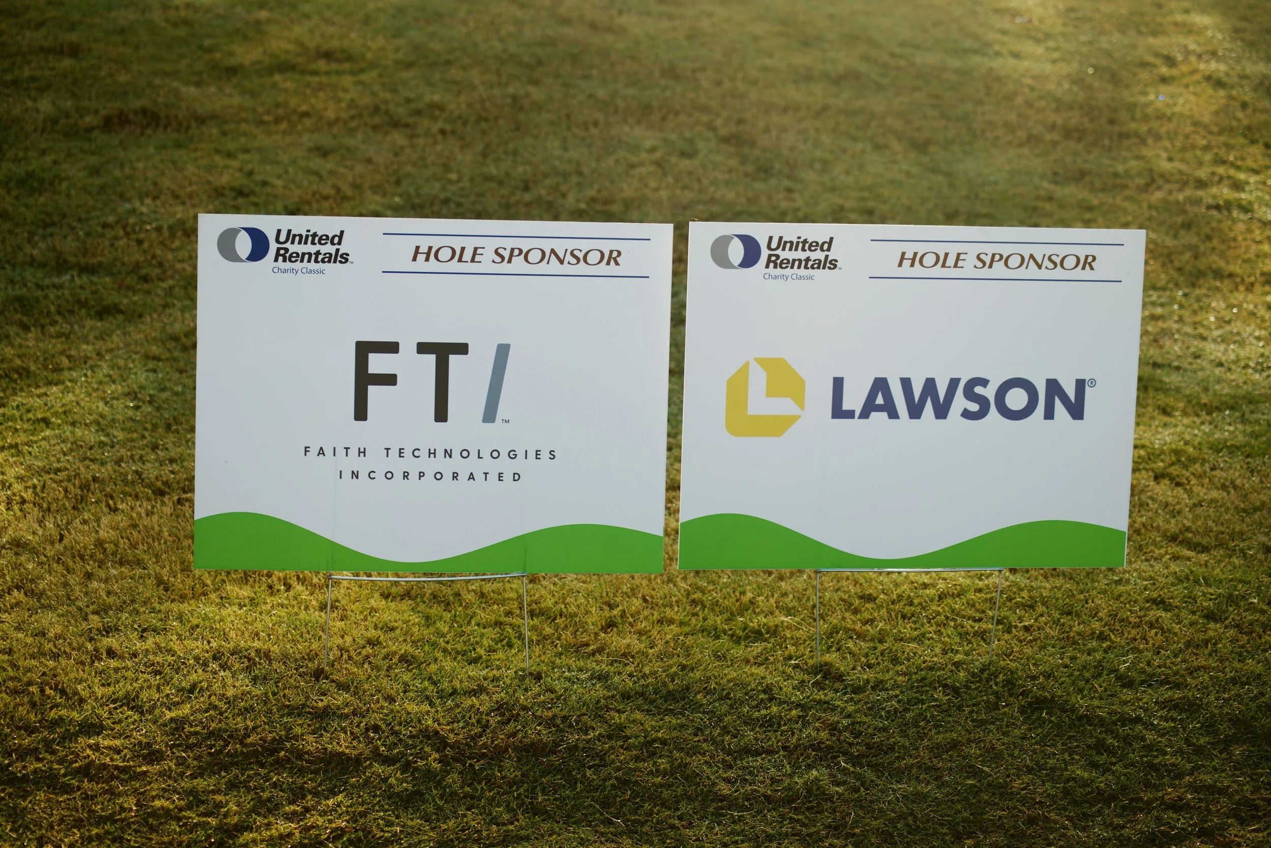 Two signs on a grassy field, the left one reads "FT Faith Technologies Incorporated," and the right one reads "Lawson" with the words "HOLE SPONSOR" above both signs, indicating sponsorship at an event.