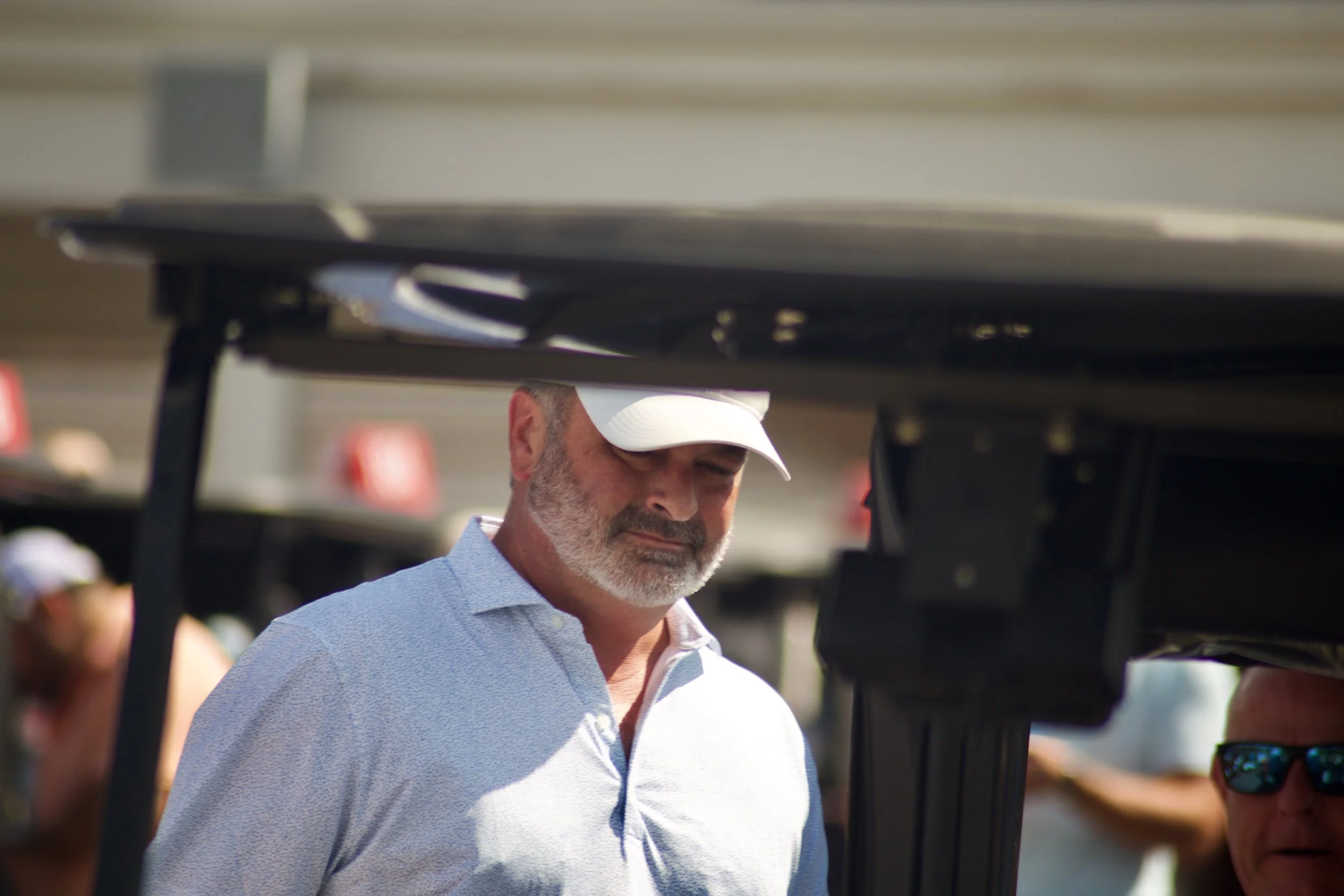 Man with gray beard and white cap looking towards the ground in a crowded outdoor setting.