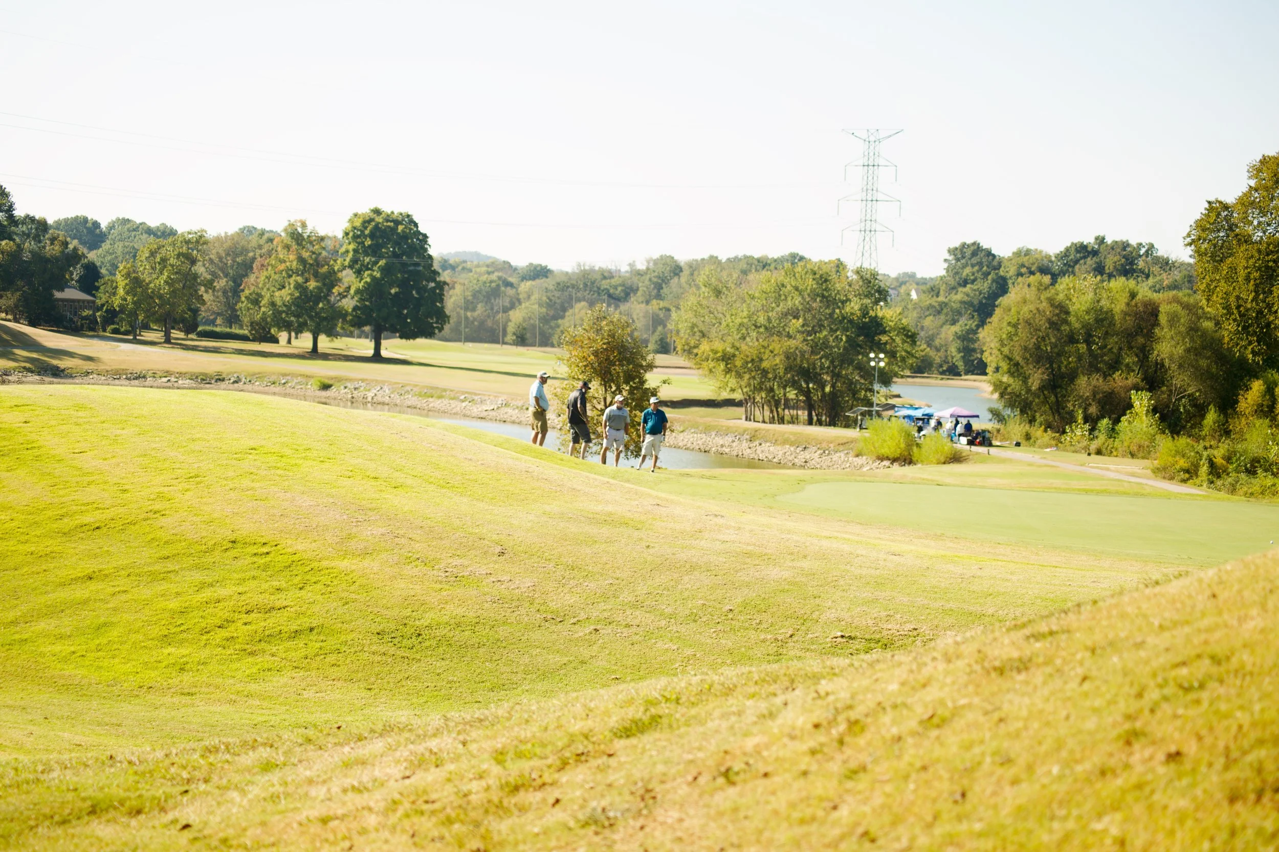 A golf course with four people walking near a water hazard, surrounded by trees and a clear sky.