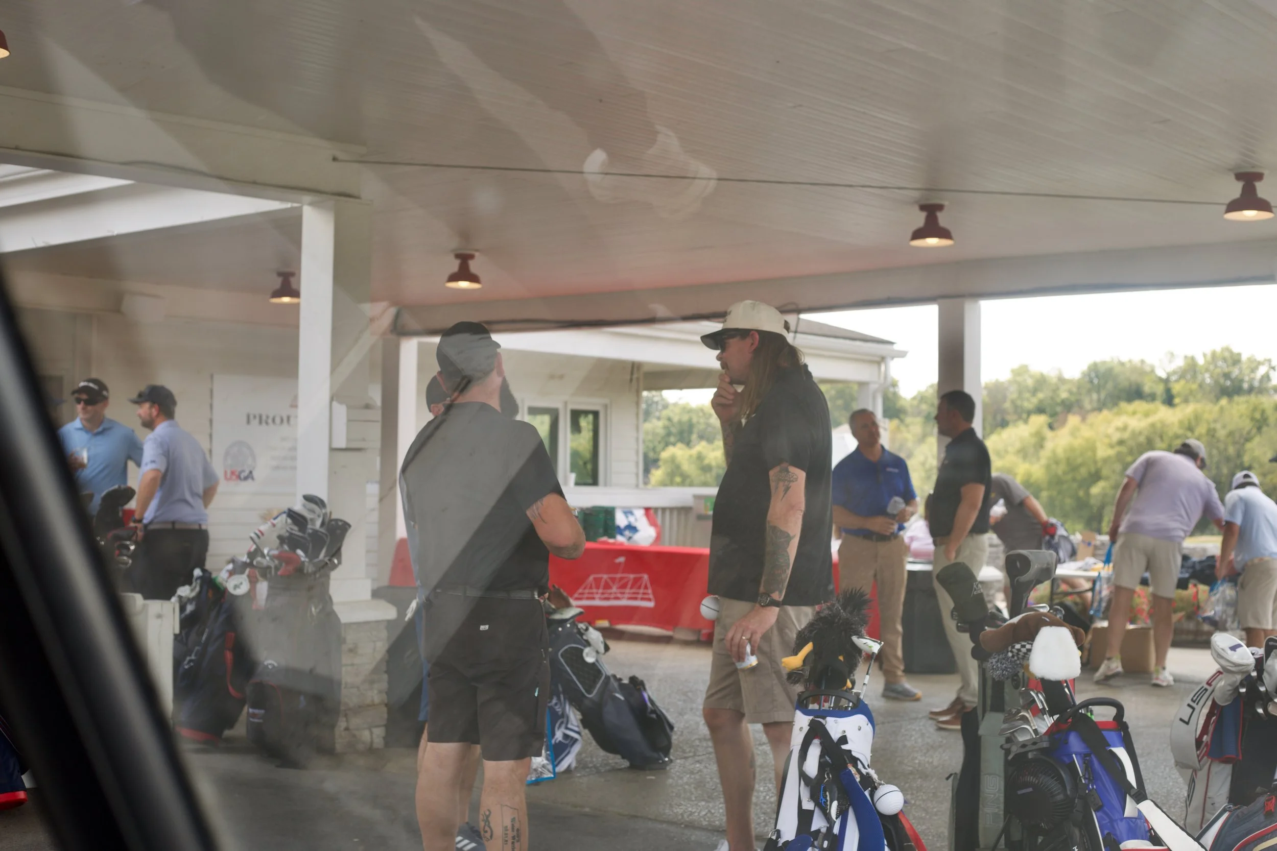 People gathering outdoors at a golf event, with golf bags and equipment visible under a covered area.