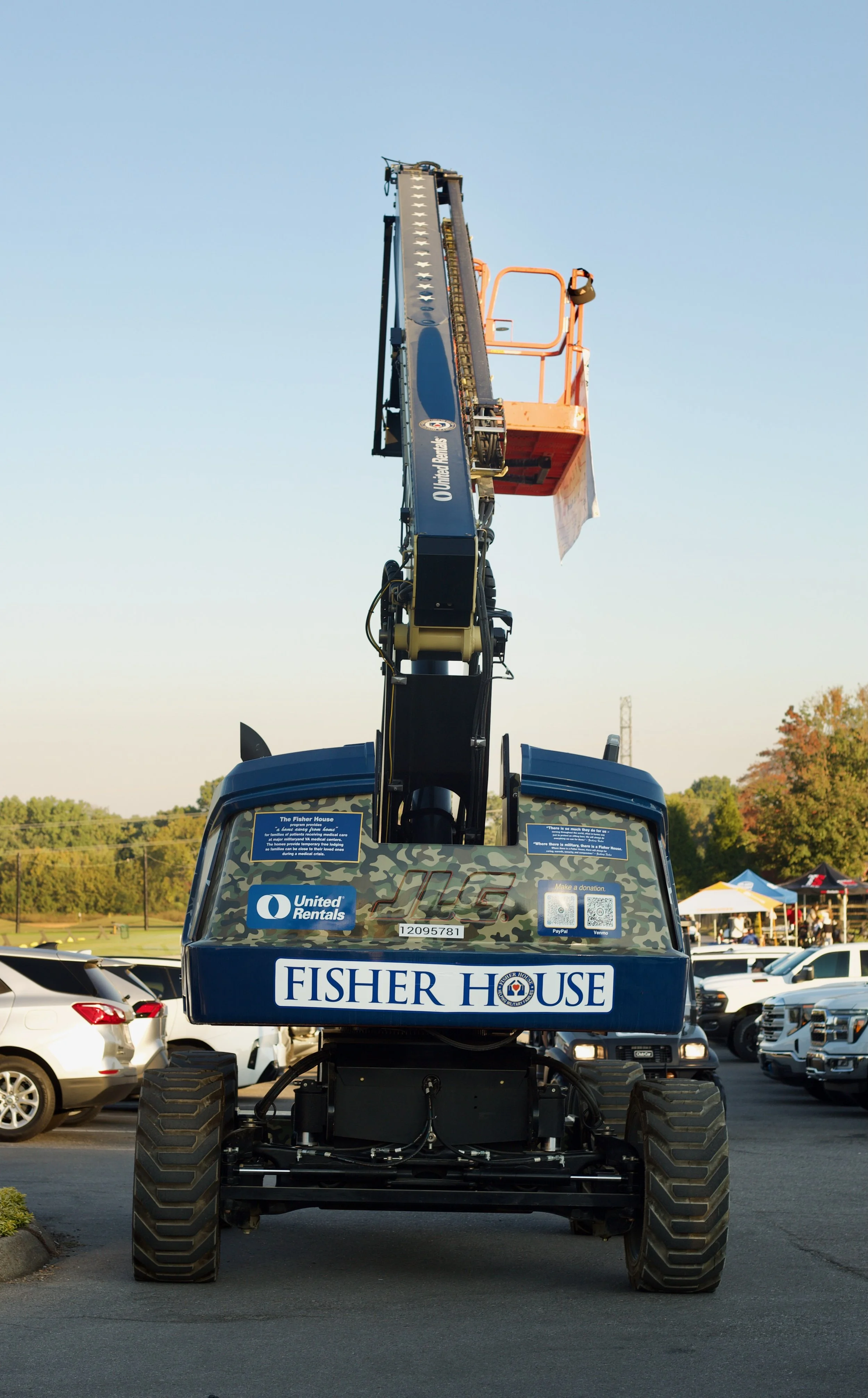 A large construction vehicle with an extended arm and bucket, branded with Fisher House and United Rentals logos, parked in a lot with other cars and tents in the background.