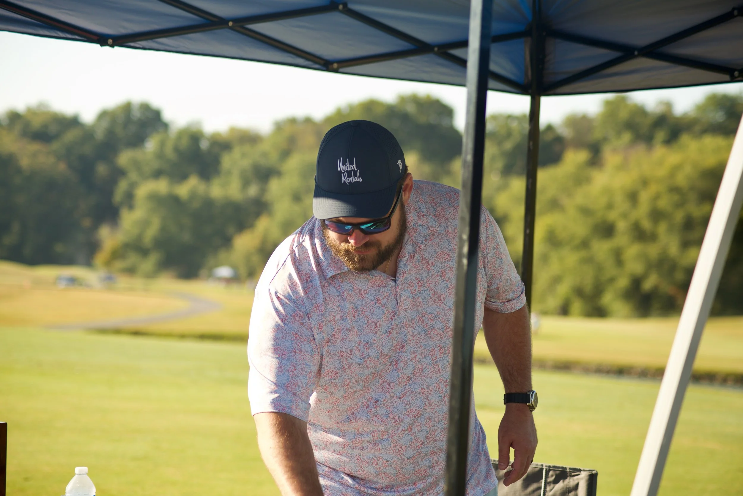 A man with a beard wearing a black cap with the logo 'United Rentals' and sunglasses, working outdoors under a blue canopy in a park or field with trees in the background.