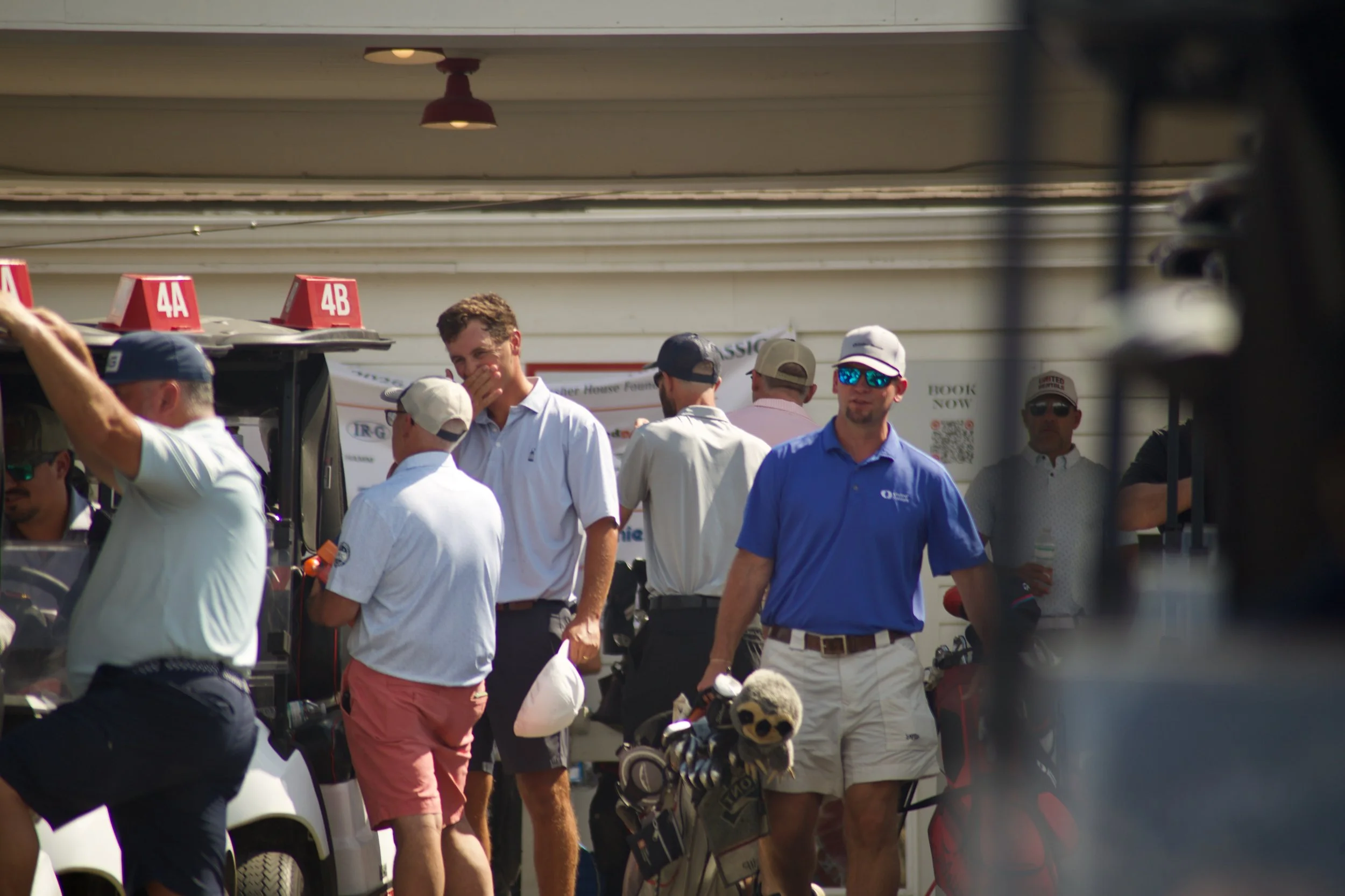 Group of men wearing casual clothes, some with hats and sunglasses, gathered outdoors near golf carts and golf bags, possibly at a golf course or event.