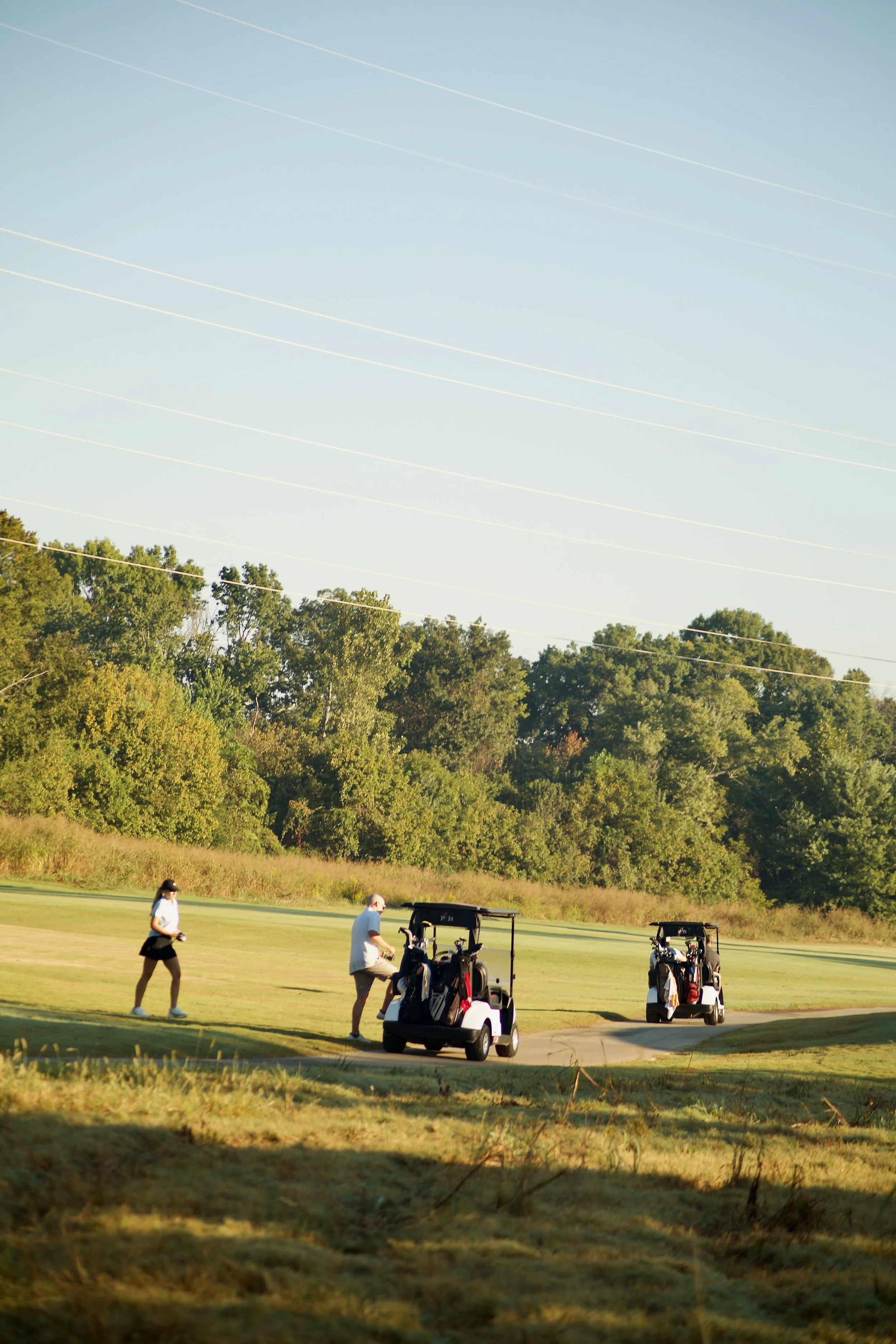 Two women and two men on a golf course with golf carts, trees in the background, and a clear sky overhead.