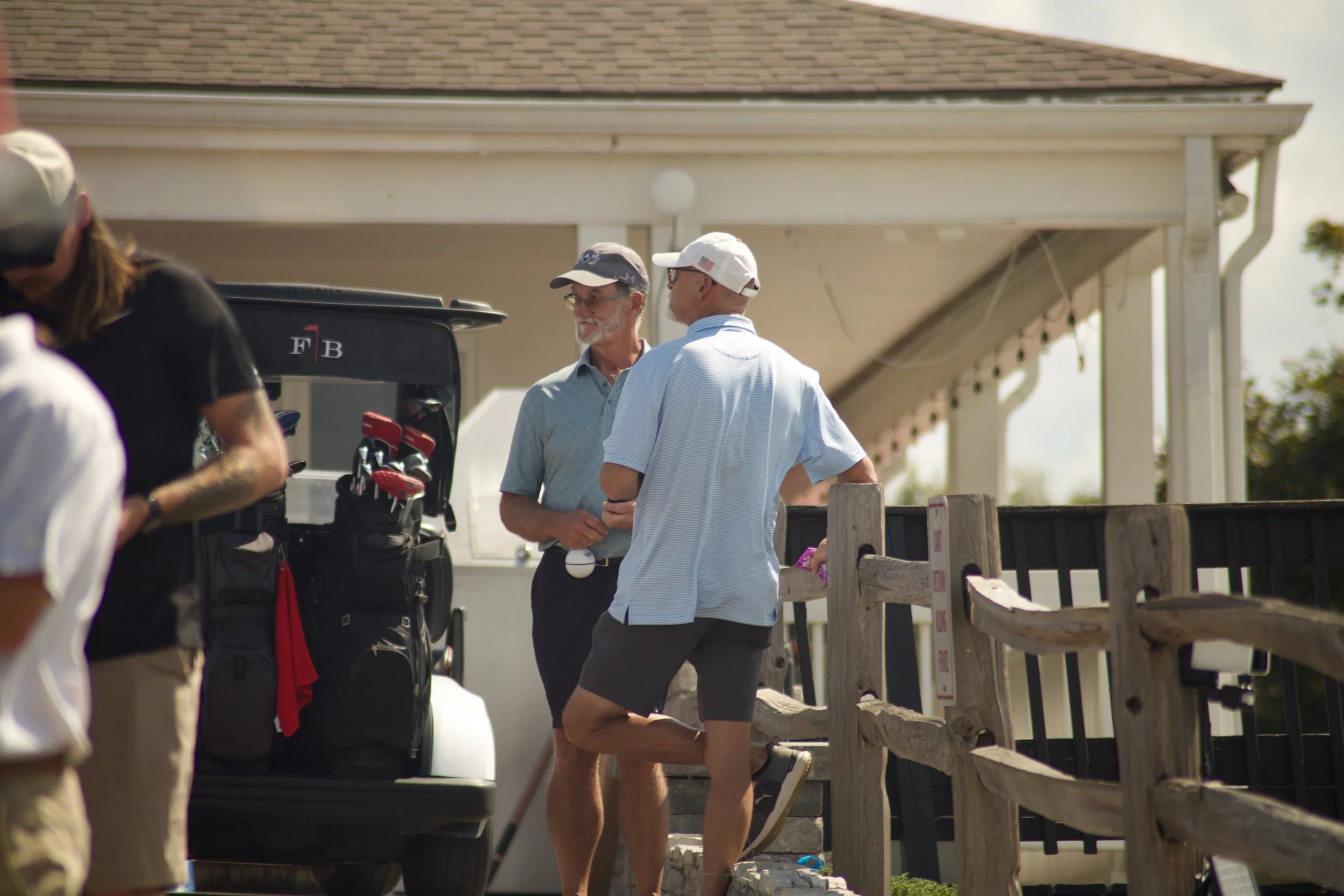 Two men conversing outdoors near a wooden railing, one wearing a light blue polo shirt, dark shorts, and a cap, the other in a light blue striped shirt, dark shorts, and a white cap, with golf clubs nearby.