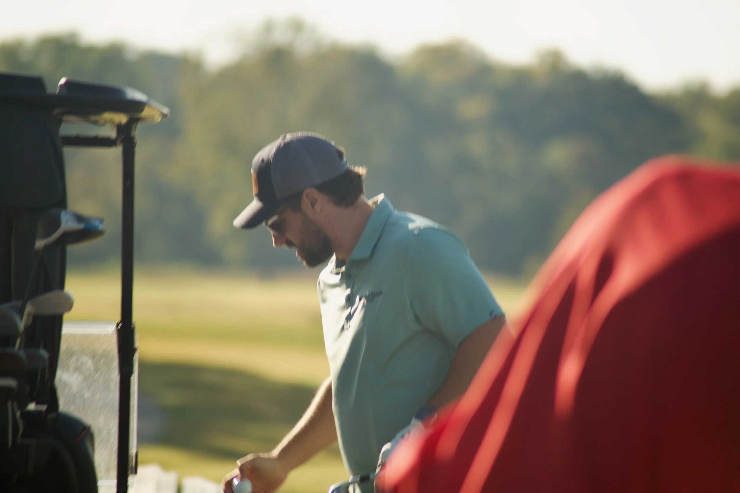 A man wearing a baseball cap and sunglasses adjusting a golf club on a golf course, with trees and a blue sky in the background.