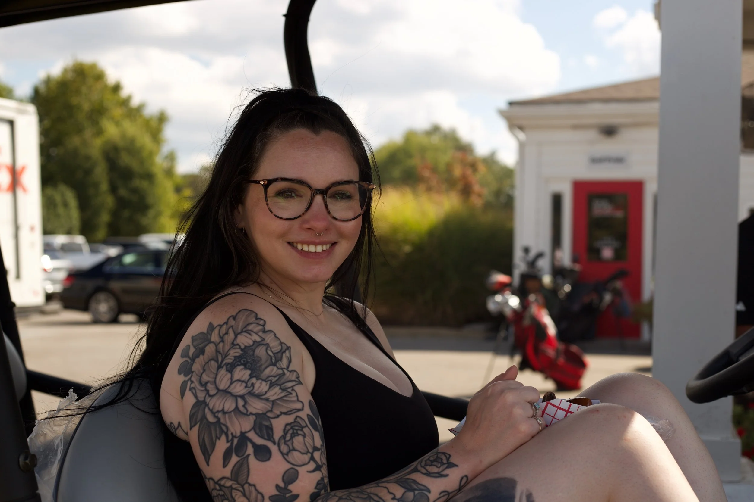 A woman with glasses and tattoos smiling while sitting outdoors in a golf cart.
