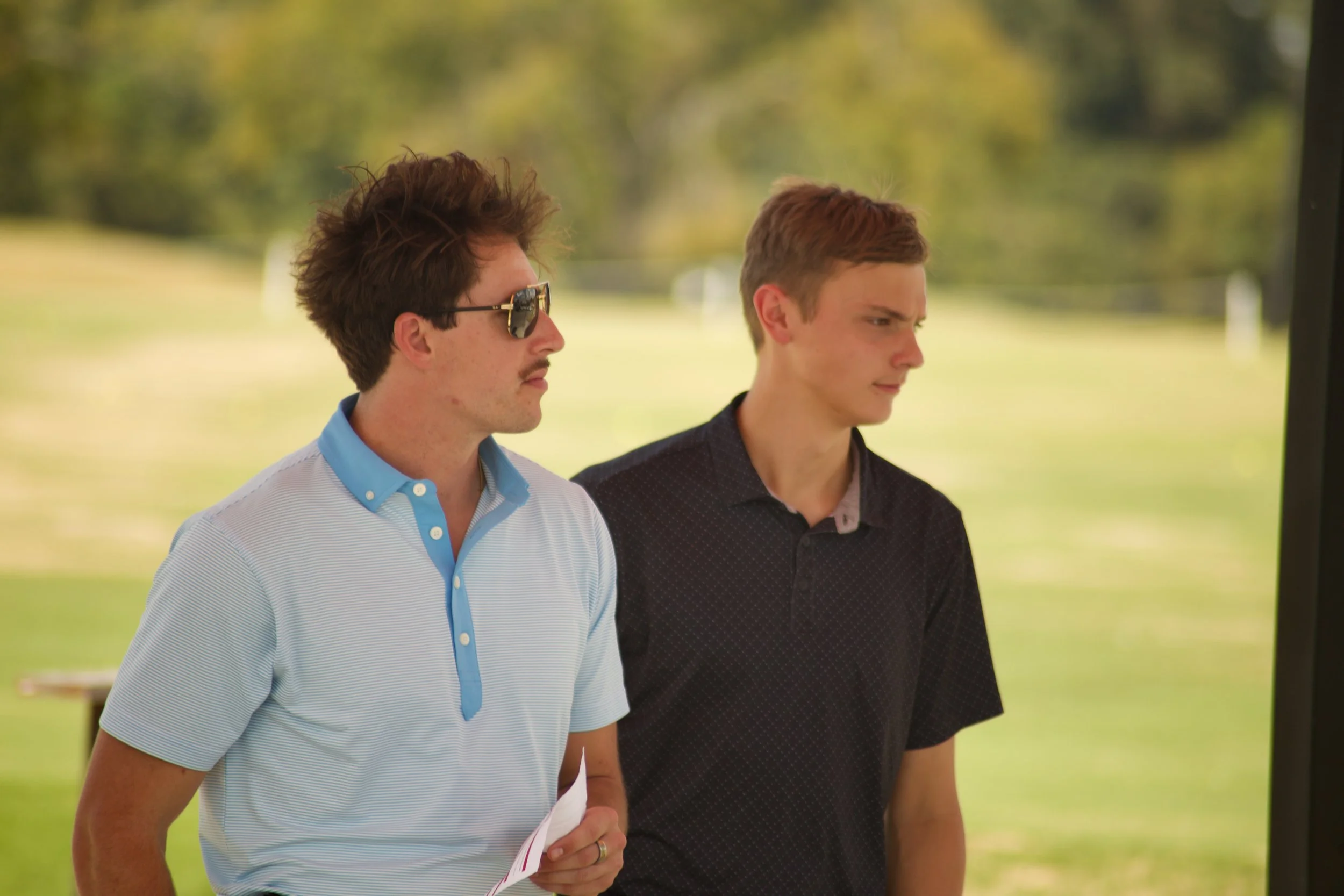 Two young men standing outdoors near a black object, possibly a screen, with blurred green trees and grass in the background, one wearing sunglasses and a light striped polo shirt, the other with short brown hair wearing a dark patterned shirt.