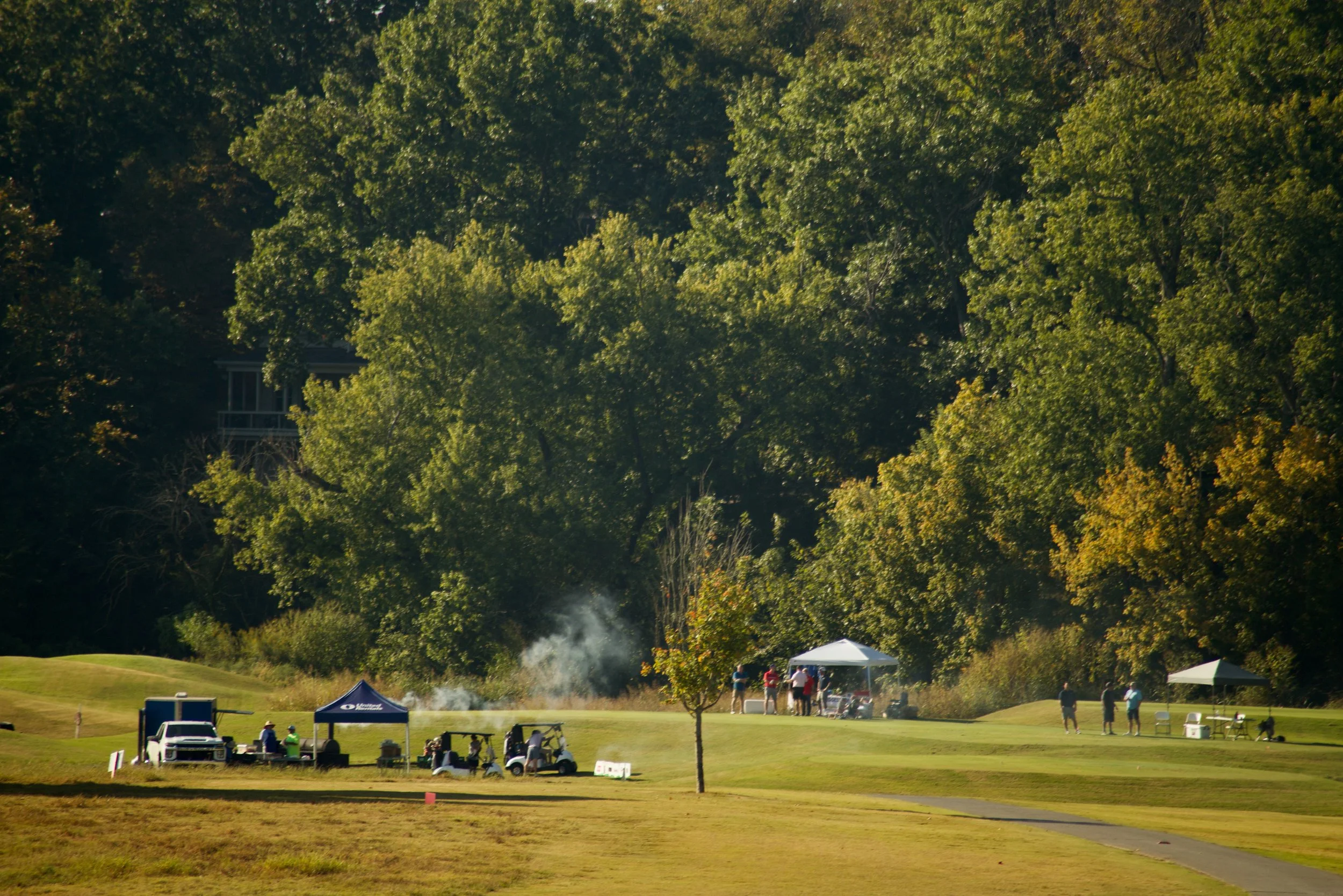 A golf course with green grass, trees, tents, and people near a small group of trees. There is a golf cart and some golf carts nearby, indicating a golf event or gathering.