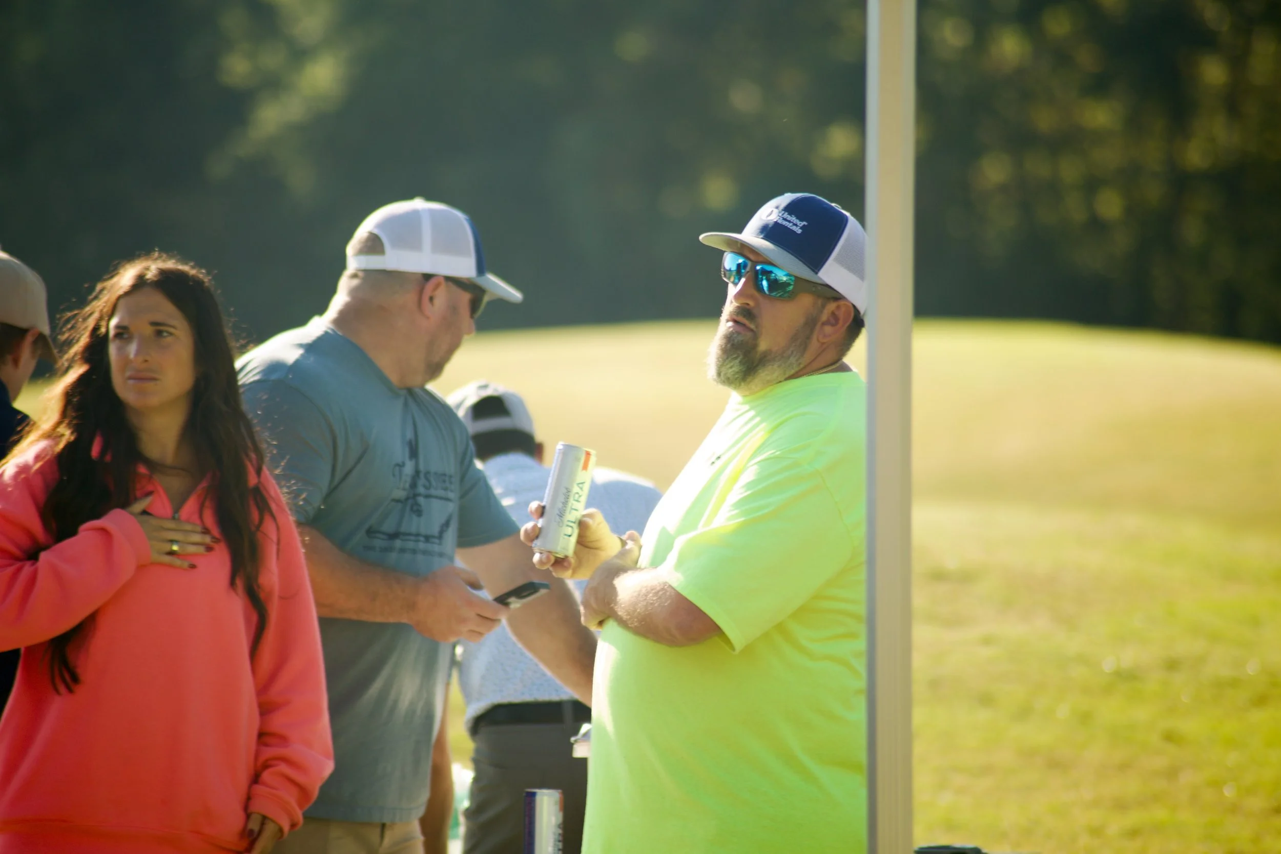 A group of people outdoors near a flagpole, with a grassy field and trees in the background. One woman in a pink jacket appears emotional, one man in sunglasses and a cap is looking at his phone, and another man in a bright yellow shirt and cap is ho