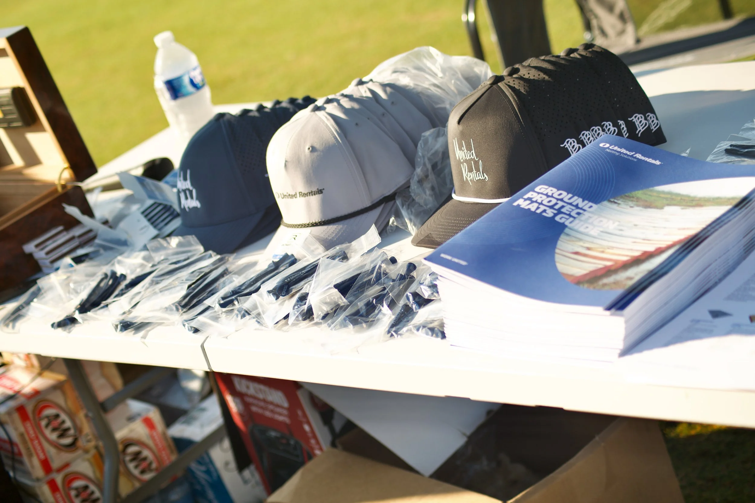 Outdoor table displaying baseball caps, brochures, and small items, with a bottle of water and a box of supplies in the background.