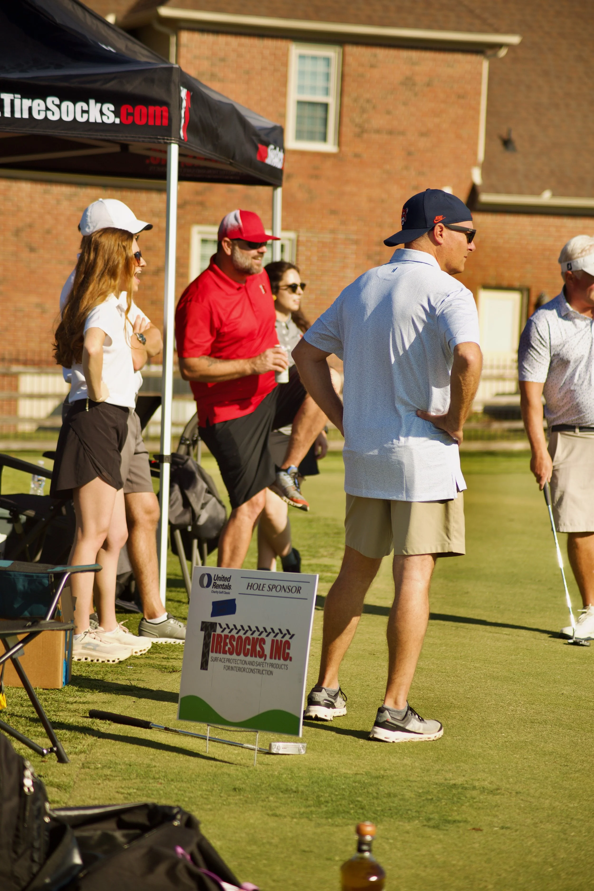 Group of people playing mini golf outdoors on a sunny day, with a golf cart and a sign reading 'Hole Sponsor' in the foreground.
