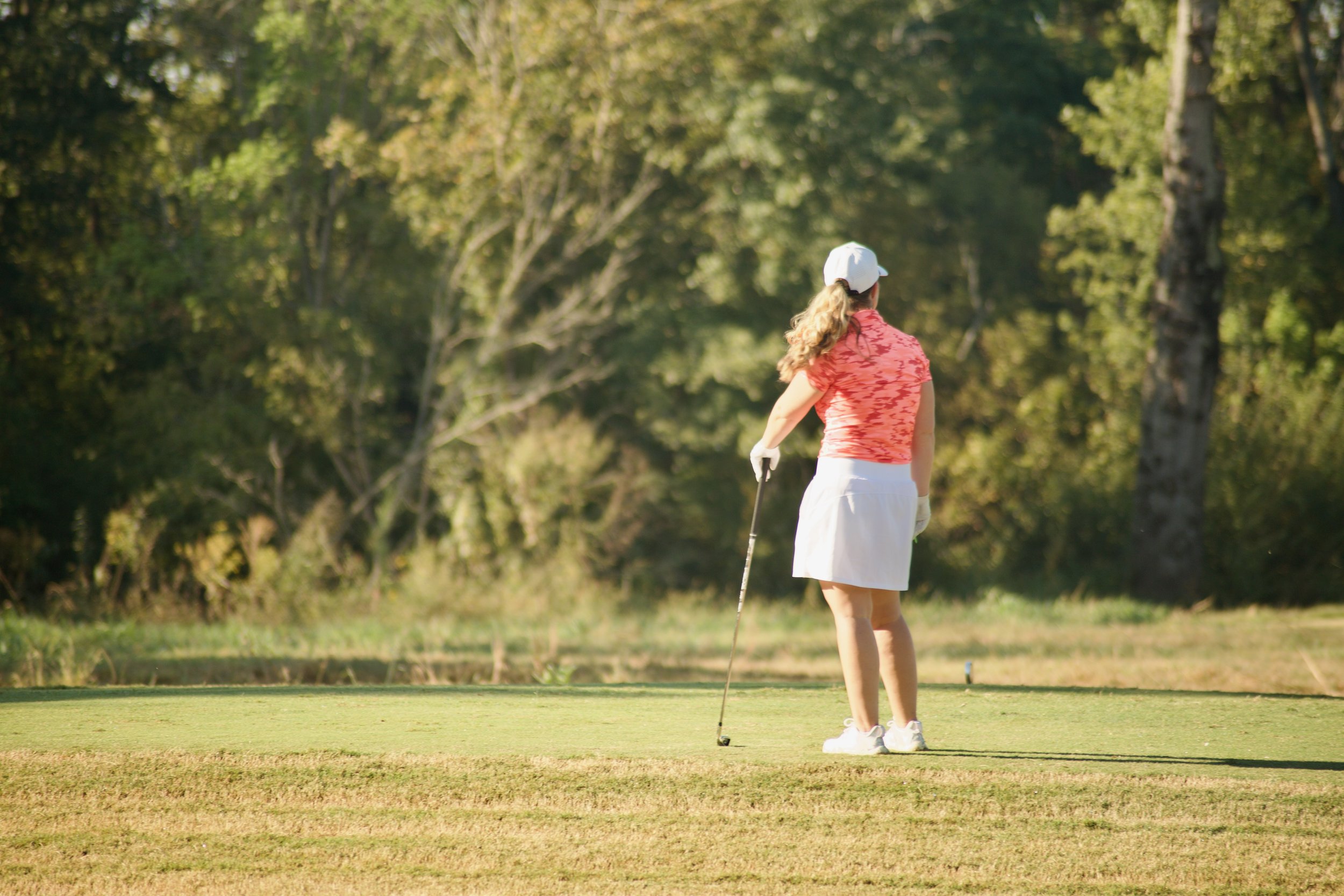 A woman with curly blonde hair wearing a white cap, coral and white patterned polo shirt, white skirt, and sneakers stands on a golf course preparing to putt, surrounded by green grass and trees.