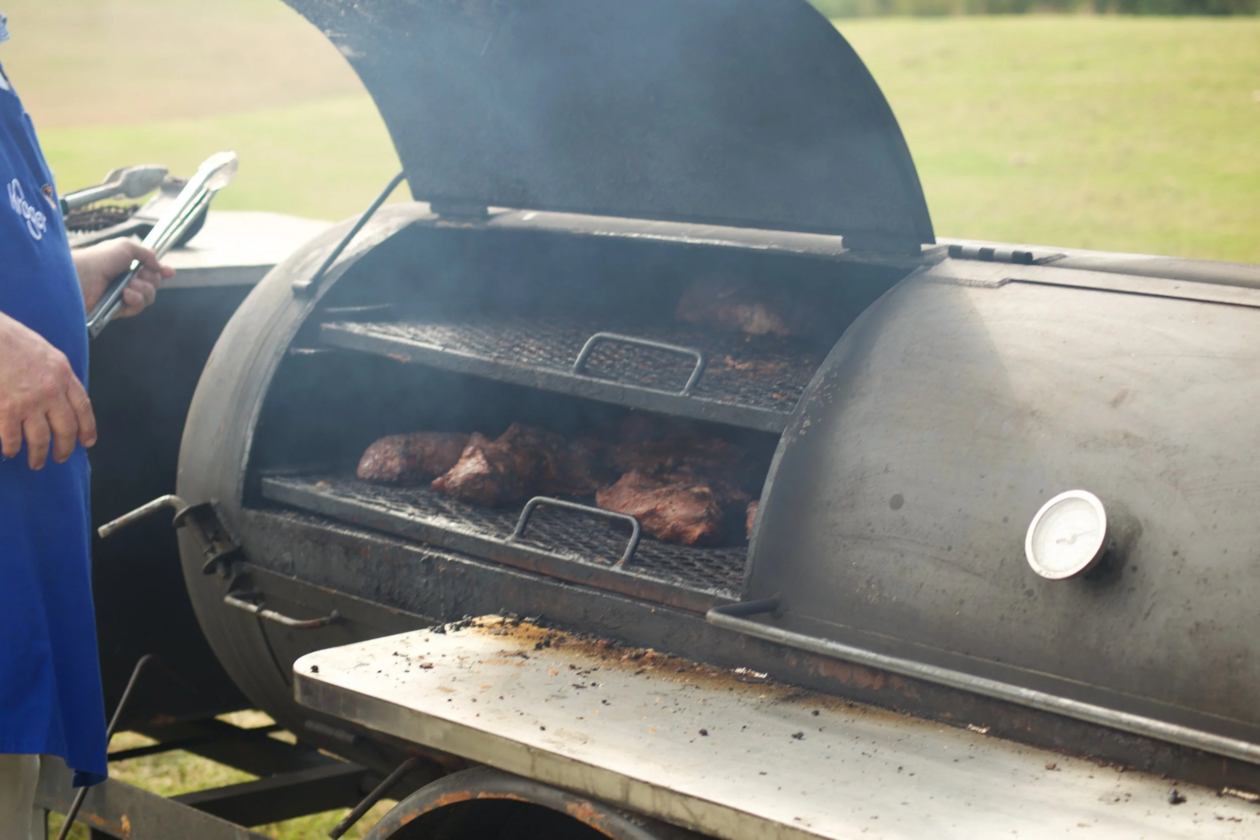 A person in a blue shirt grilling meat on a barbecue grill outdoors. The grill lid is open, showing cooked meat inside. The background is a grassy outdoor area.
