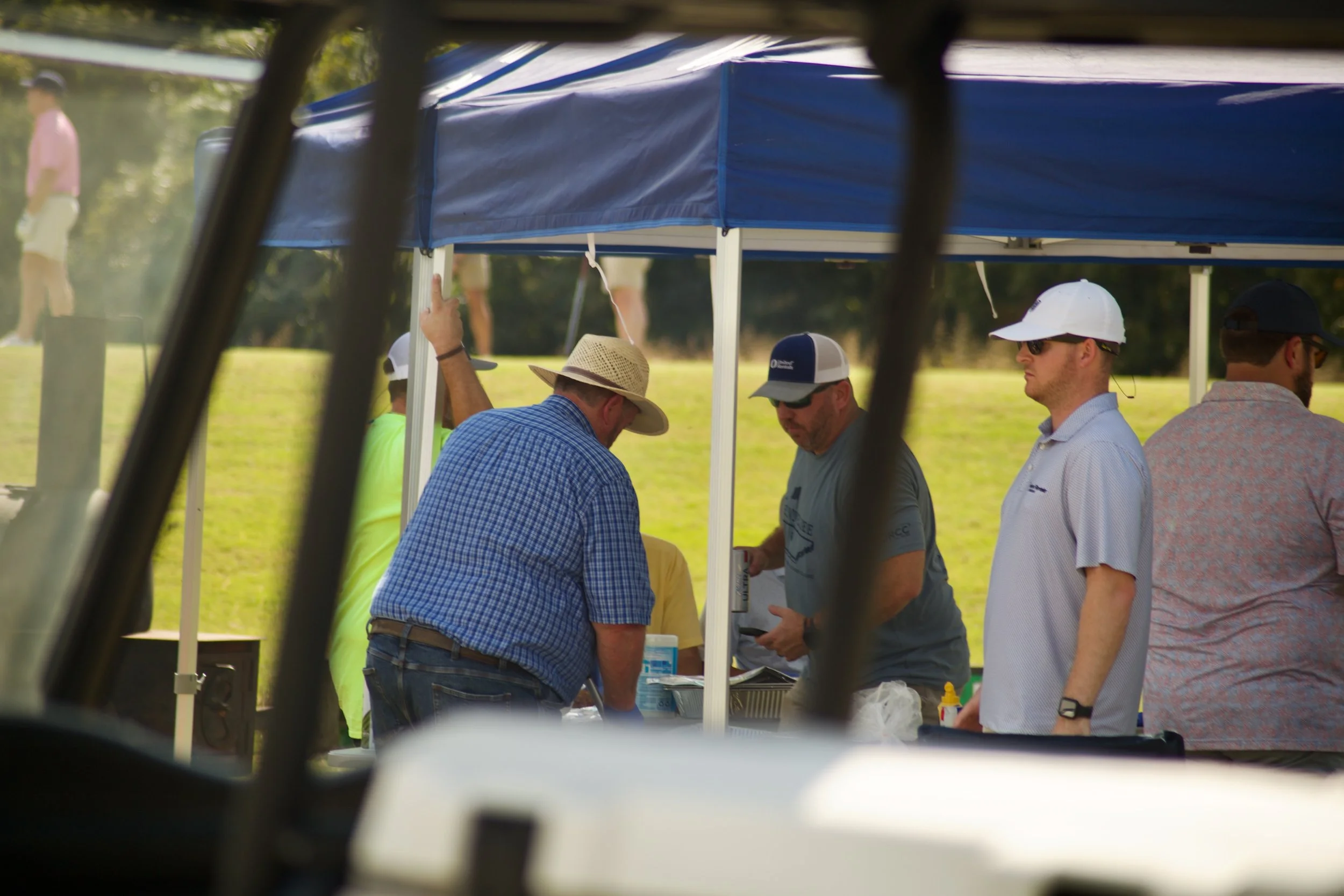 People standing under a blue canopy tent at an outdoor event, viewed through a vehicle window with black bars in the foreground.