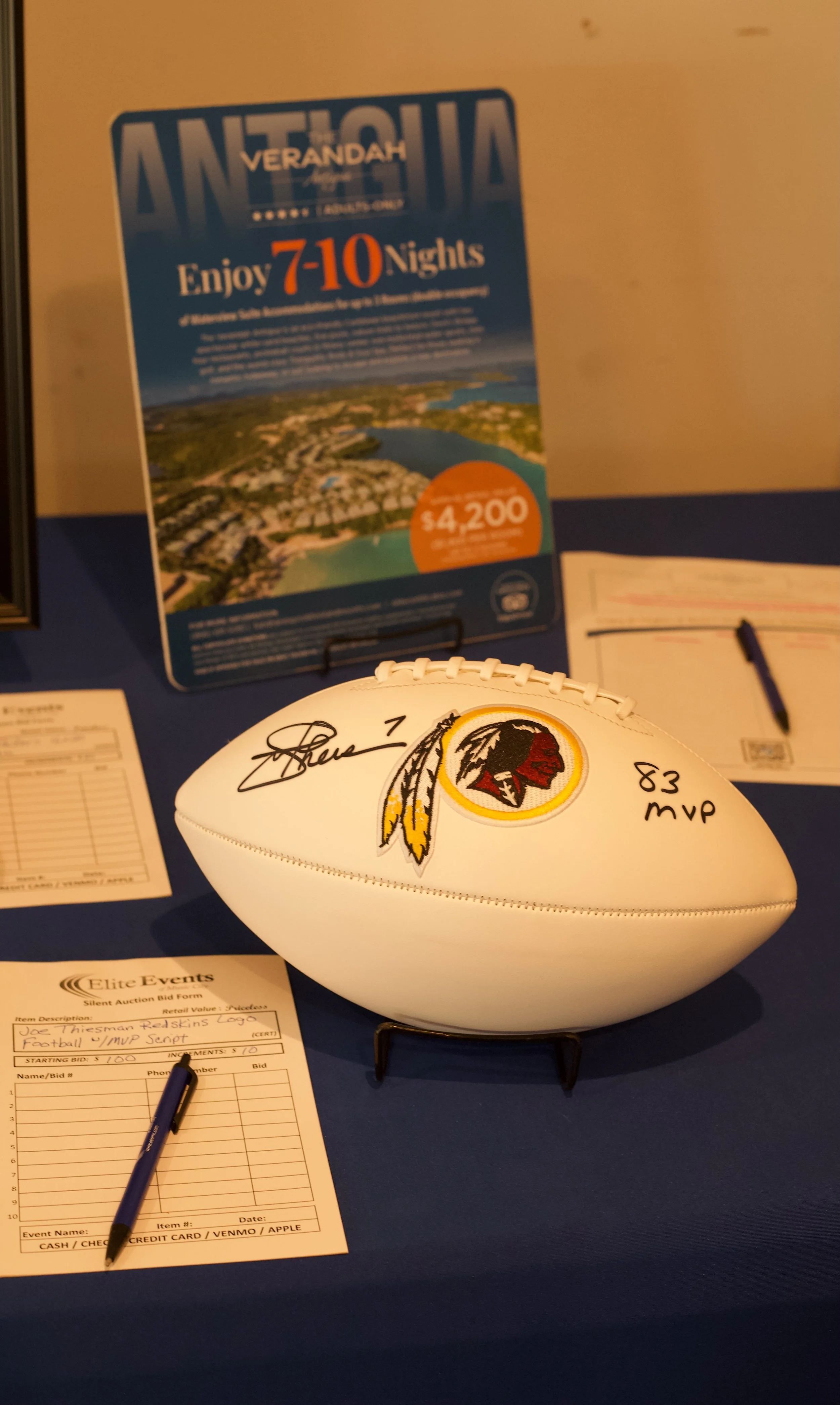 Display table with a signed football featuring a Redskins logo, an auction bid sheet, and promotional material for Antigua Verandah hotel, advertising stays of 7 to 10 nights for $4,200.