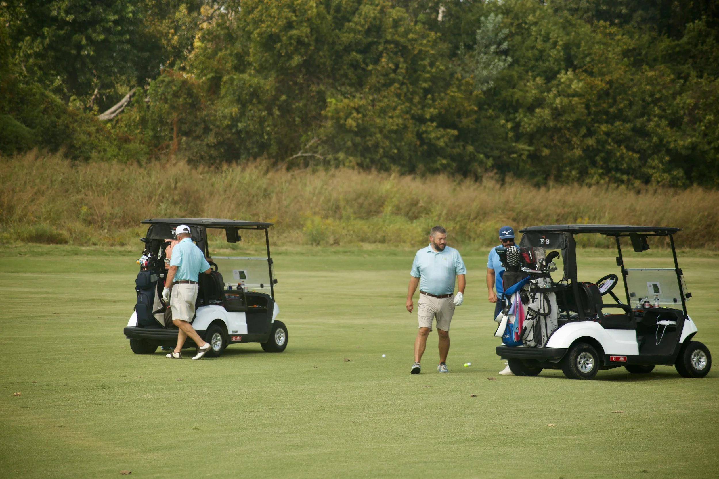 Four men on a golf course with golf carts and golf bags, surrounded by trees.