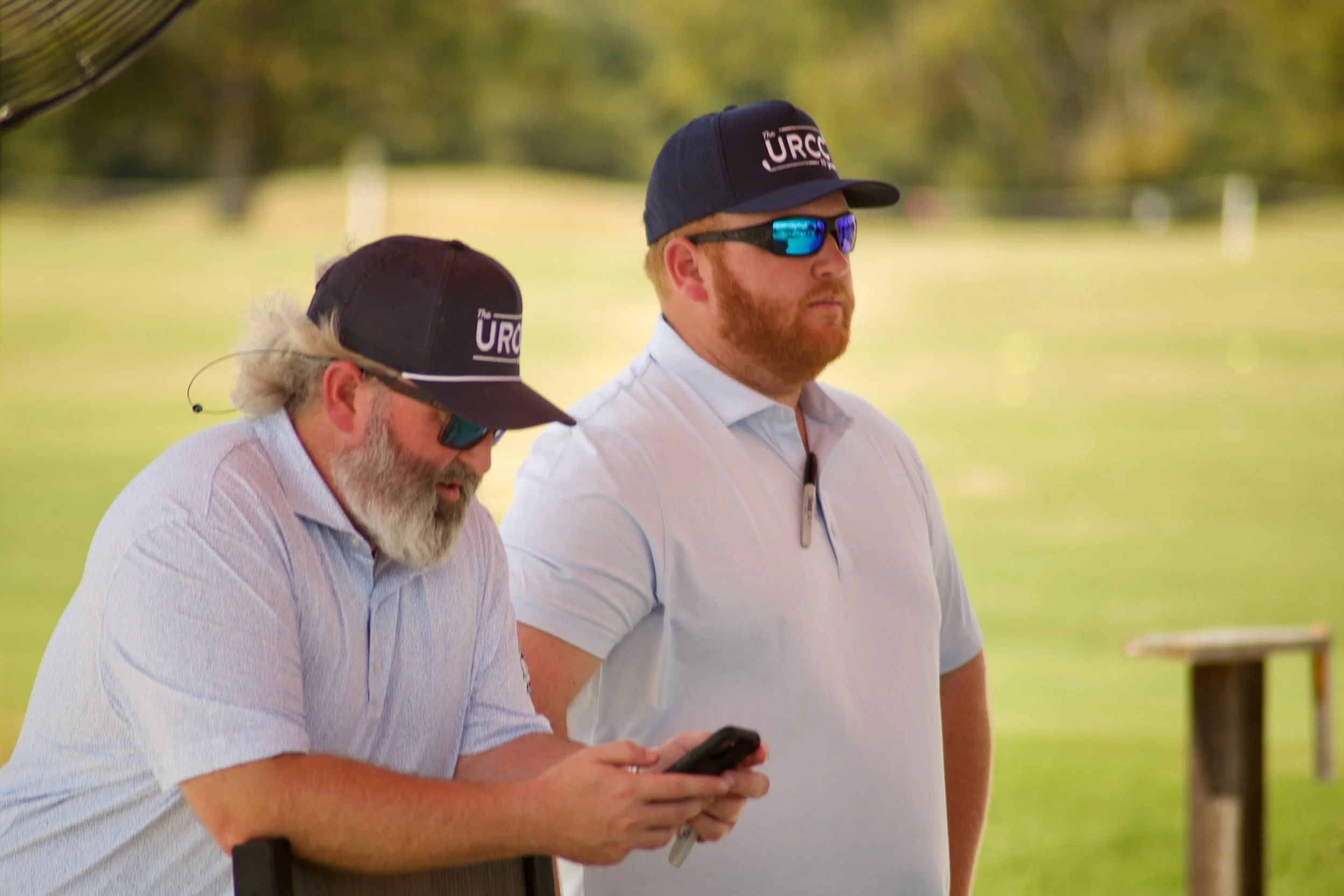 Two men wearing caps and sunglasses standing outdoors with a blurred background of green trees and grass, one is holding a phone.