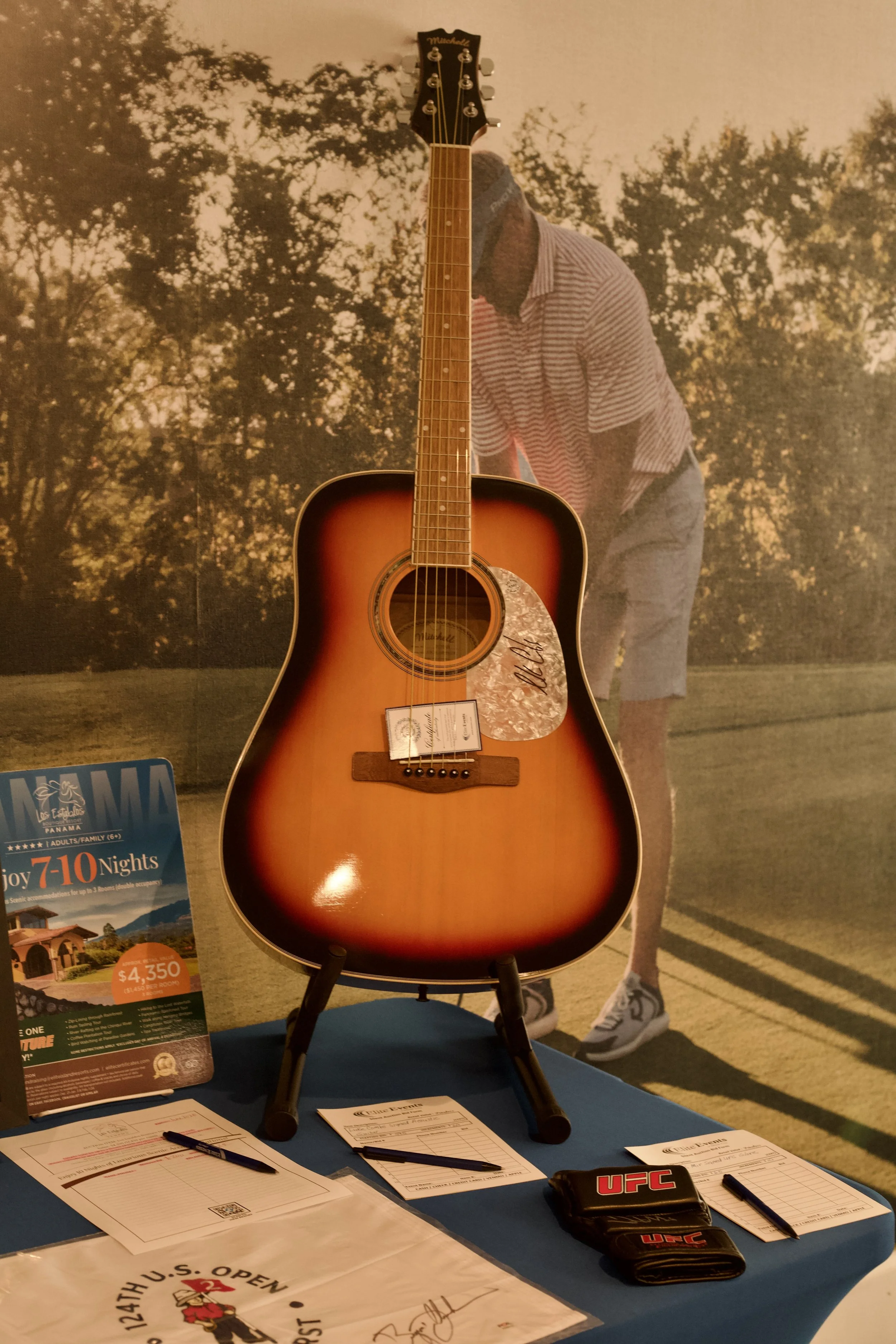 An acoustic guitar displayed on a table, with various documents, pens, and UFC branded items nearby. In the background, there is a large poster of a person in a striped shirt and shorts, with trees and sky in the background.