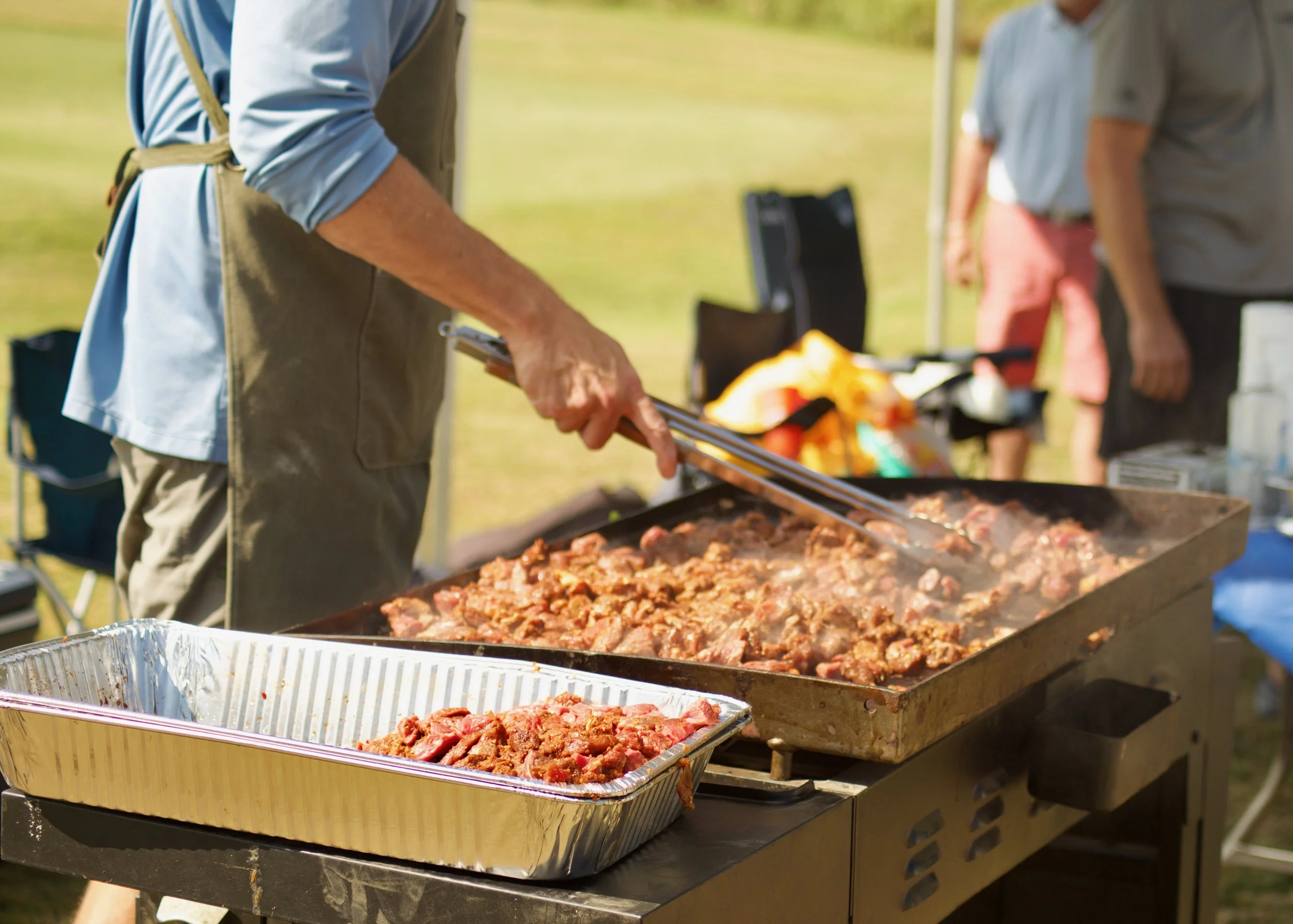 A person using tongs to serve cooked meat from a grill at an outdoor gathering, with other people visible in the background.
