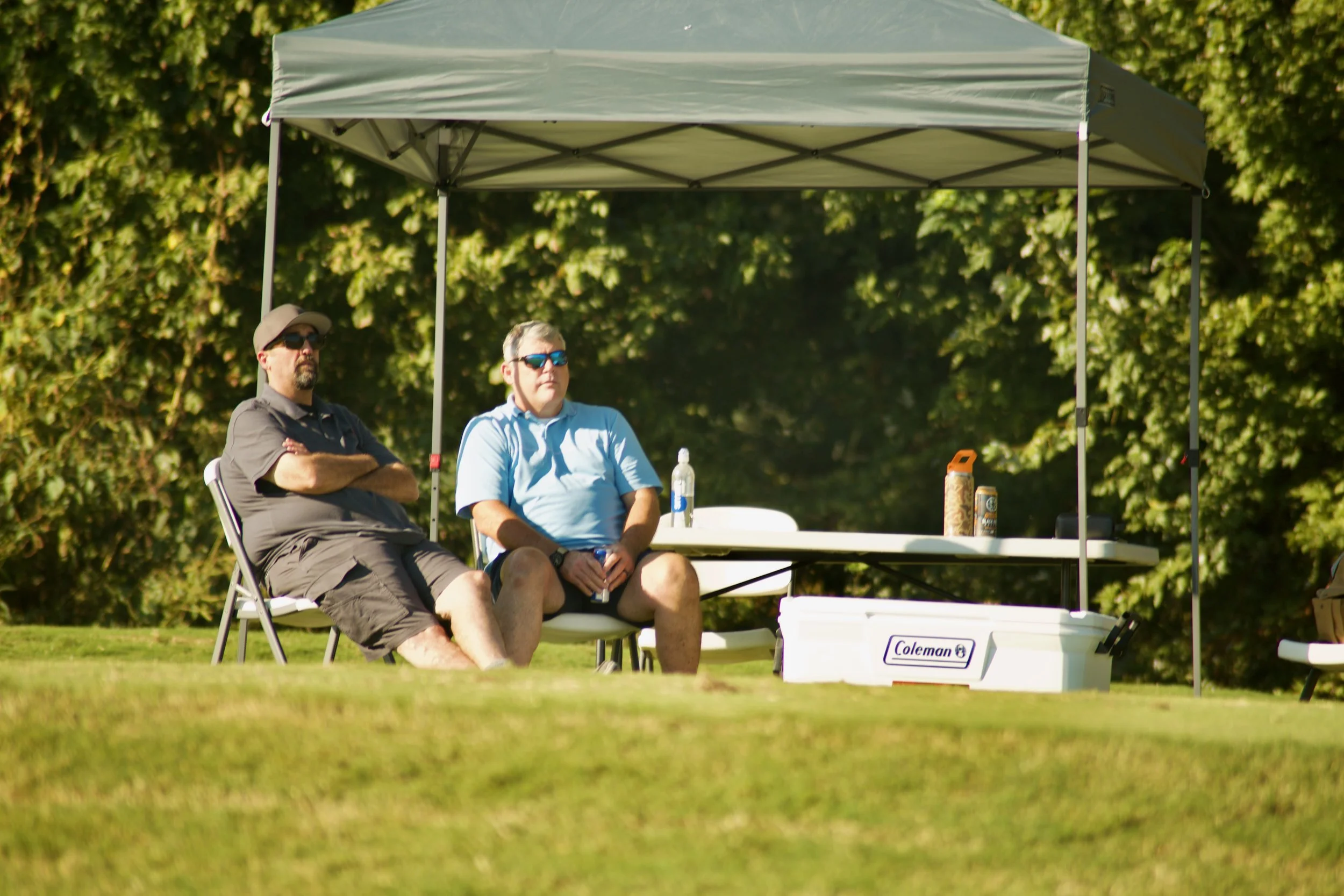 Two men sitting under a canopy on a grassy area. One man is wearing a gray shirt, shorts, sunglasses, and a hat. The other man is wearing a light blue shirt, shorts, and sunglasses. There are drinks and a cooler nearby.