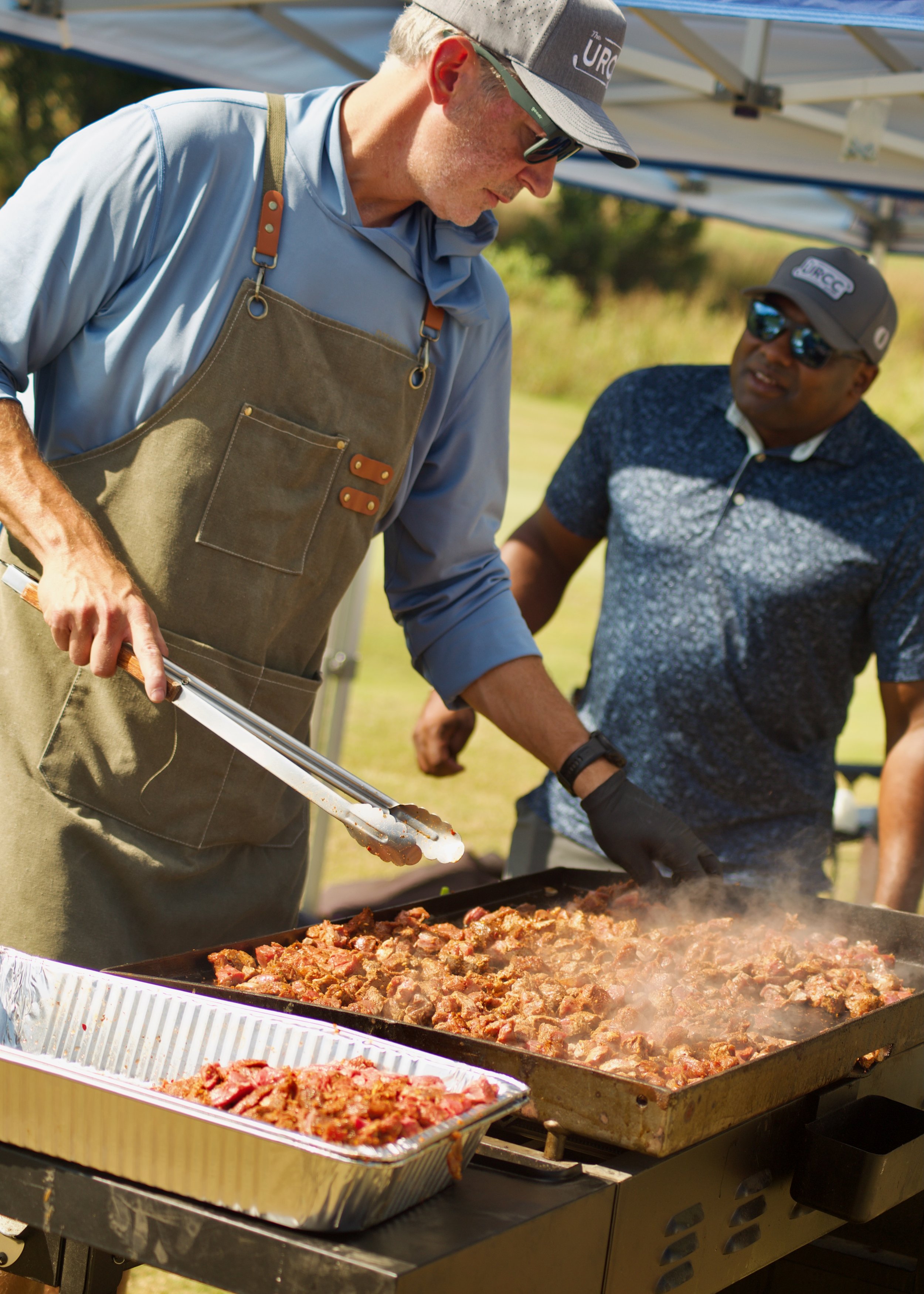 Two men are cooking and serving barbecue meat at an outdoor event. One man, wearing a brown apron and sunglasses, is using tongs to handle the meat. The other man, wearing a cap sunglasses, is standing nearby. There are large trays filled with cooked