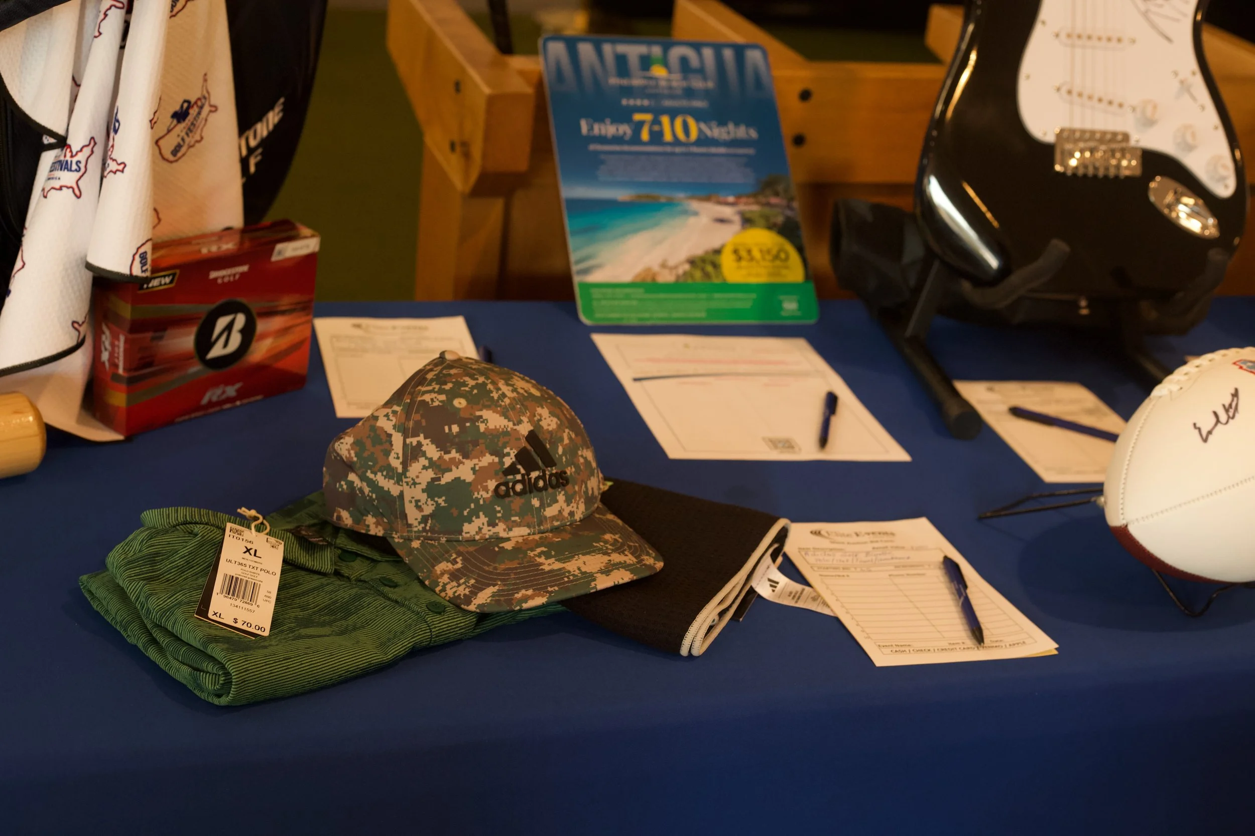 Display table with promotional materials, a camouflage patterned baseball cap, green folded clothing with a price tag, a black and white guitar helmet, and some paper forms and pens, in a retail or promotional setting.