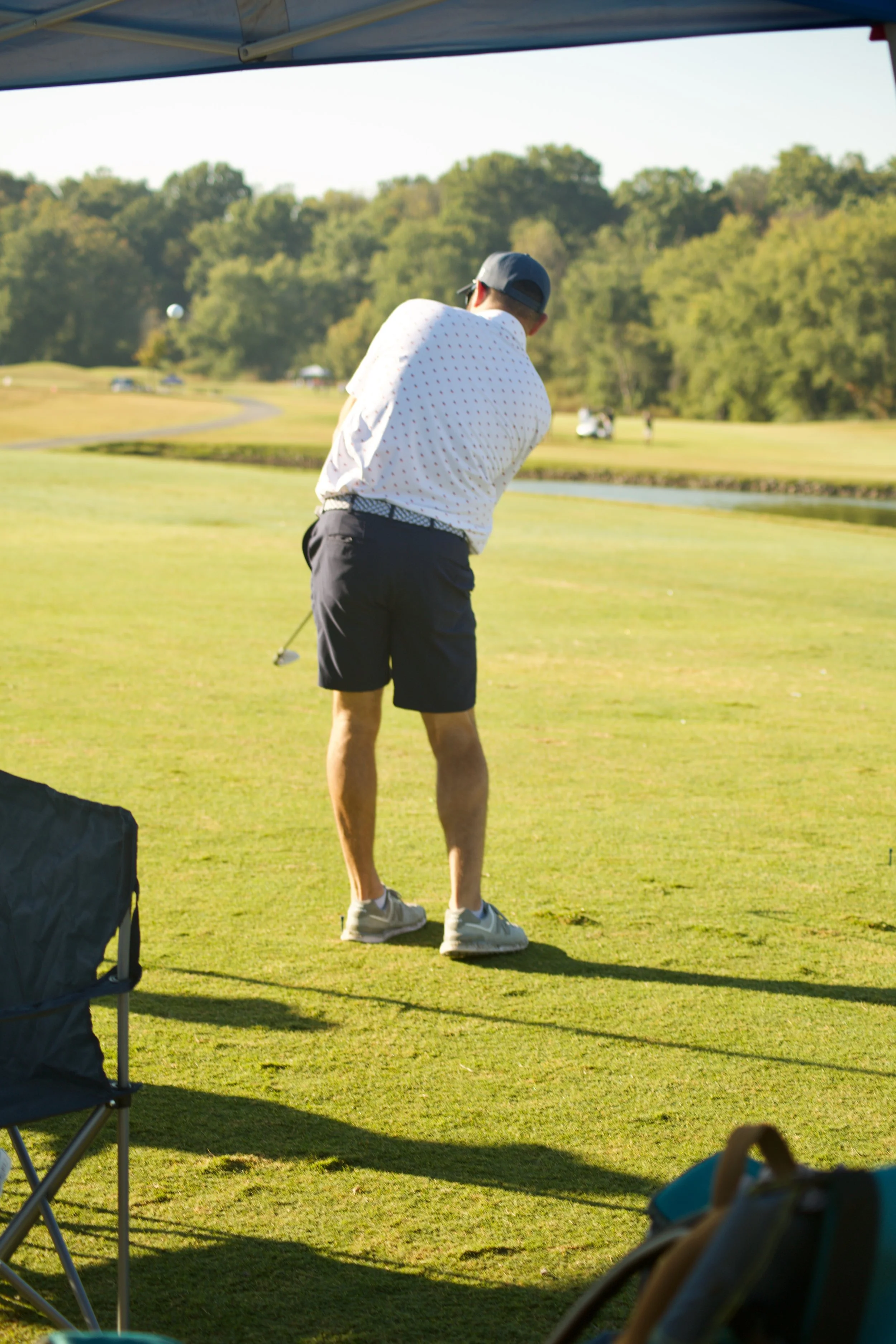 A man playing golf on a lush green golf course during daytime, wearing a white shirt, dark shorts, a cap, and sunglasses, with trees and a pond in the background.