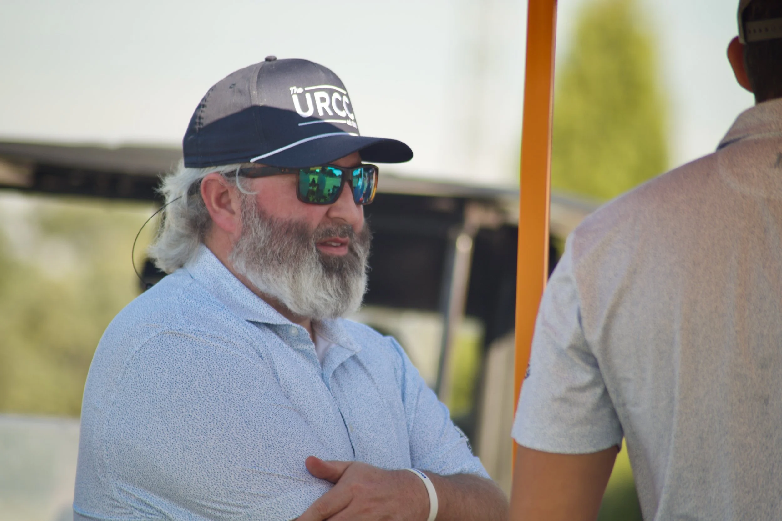 A man with a white beard, sunglasses, and a blue shirt shaking hands with another person at an outdoor event.
