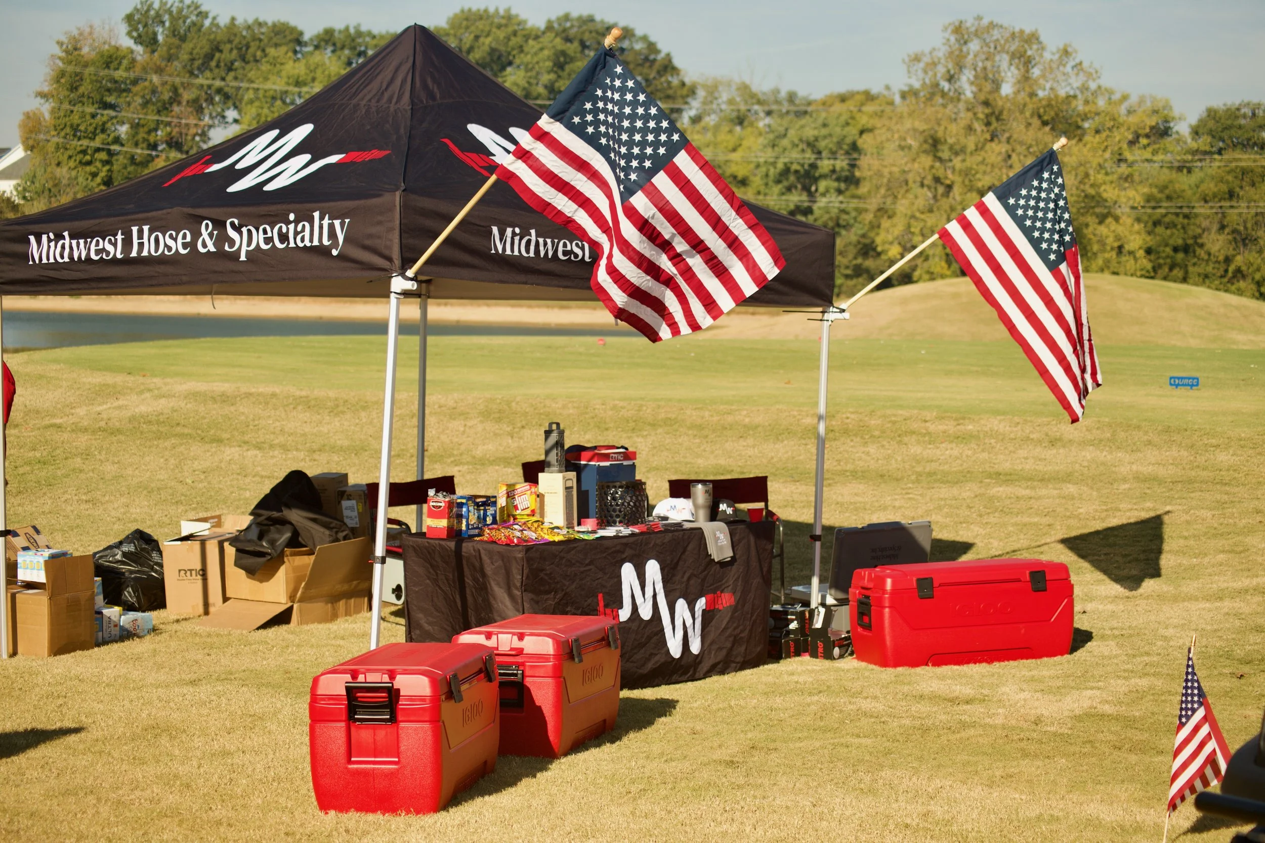 An outdoor scene featuring a black canopy tent with the words 'Midwest Hose & Specialty' and 'Midwest' printed on it. Several American flags are attached to the tent, waving in the breeze. On the ground beneath the tent, there are various boxes, bags