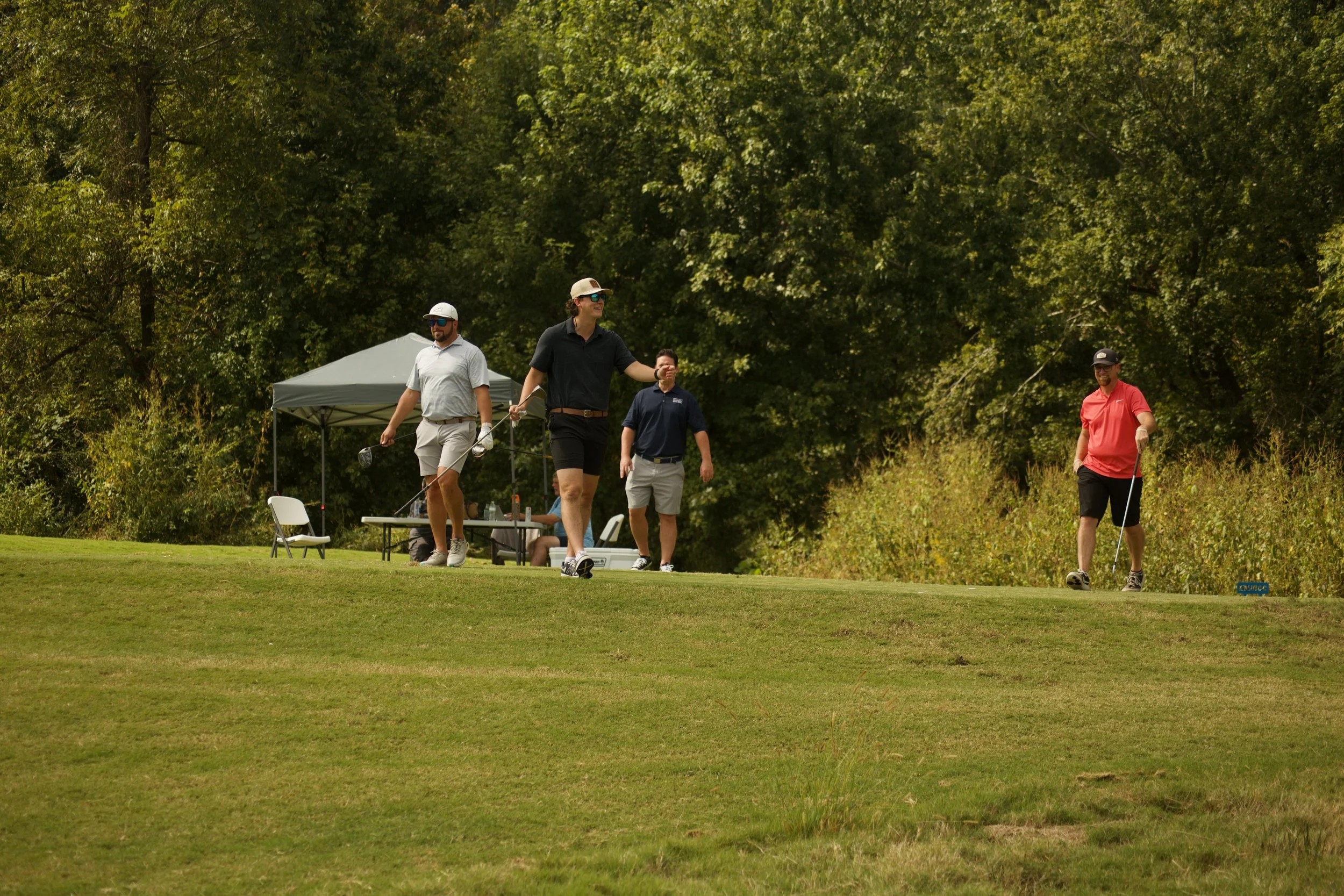 Group of four men playing golf on a course near trees, with one man putting on the green and others watching.