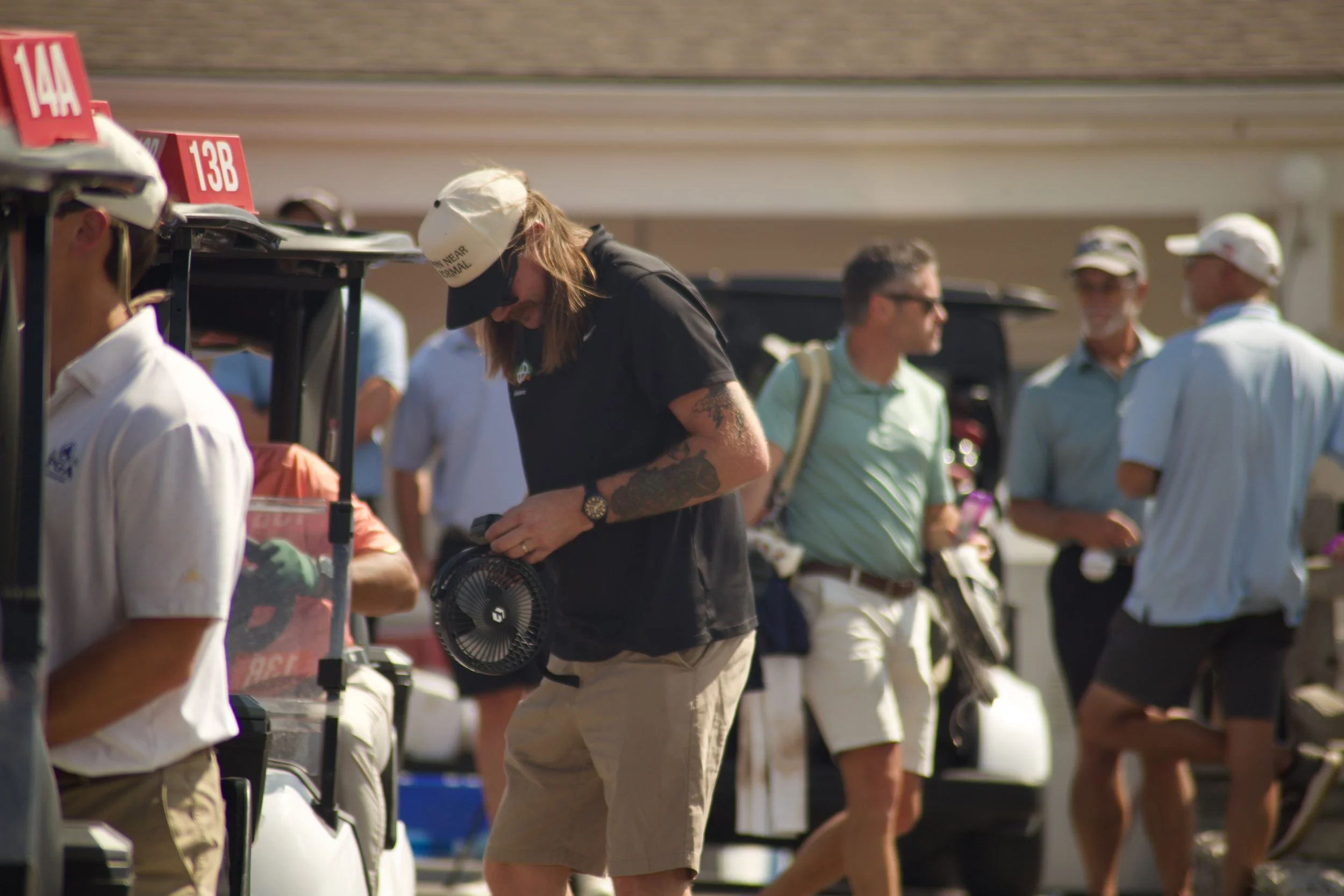 People standing in line at a golf course, some wearing caps, and a woman with tattoos holding a portable fan.