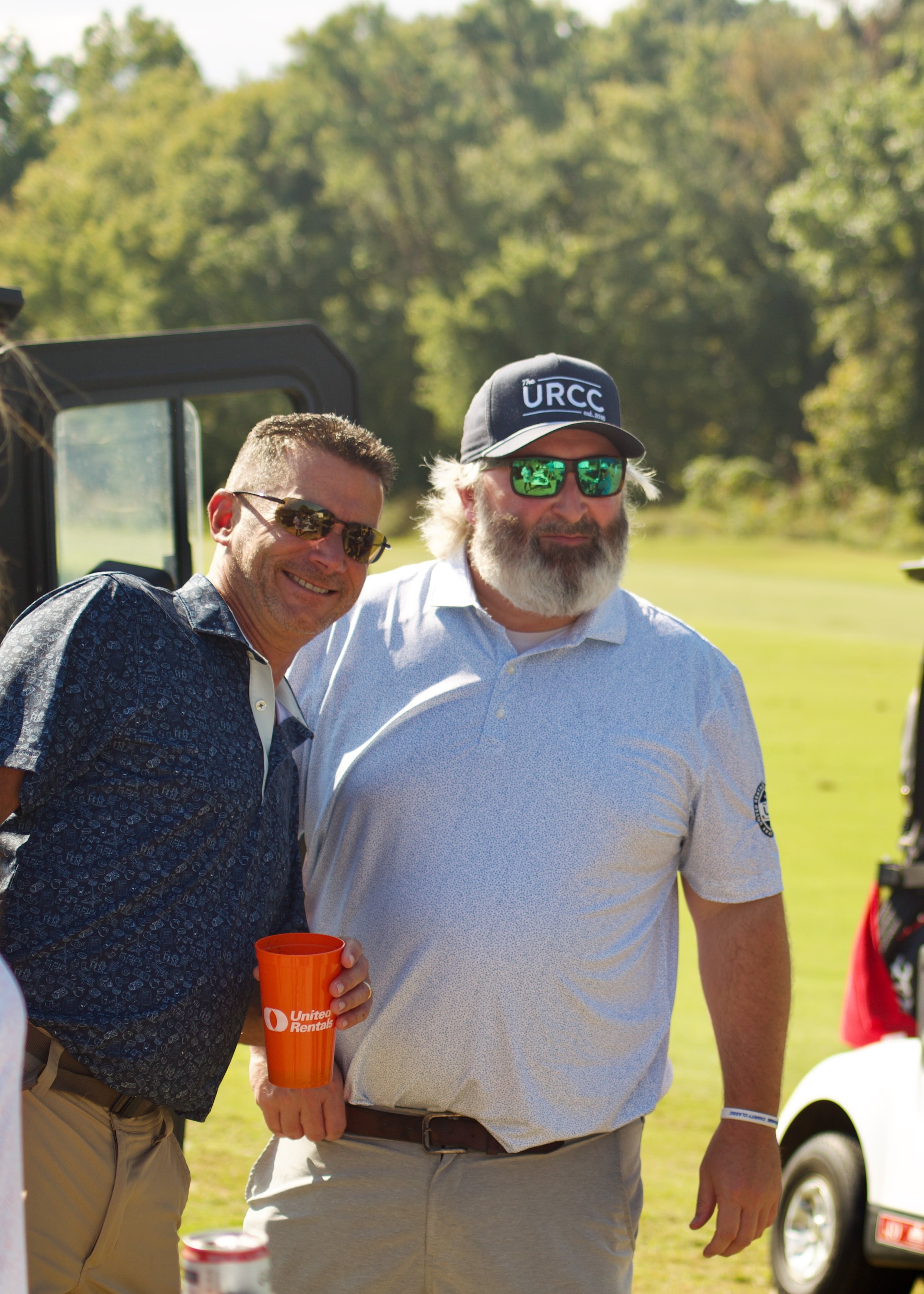 Two men standing outdoors at a sunny event, one with dark hair and sunglasses, the other with white hair, beard, and a cap that says 'URCC'. The man with white hair is wearing a light-colored polo shirt, and the other man is holding an orange cup wit