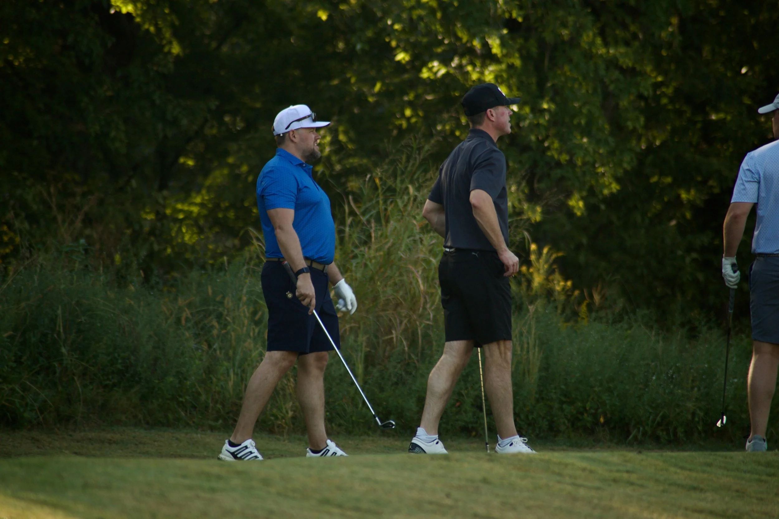 Group of men on a golf course, wearing athletic clothing and hats, standing on the grass surrounded by trees, with one holding a golf club.