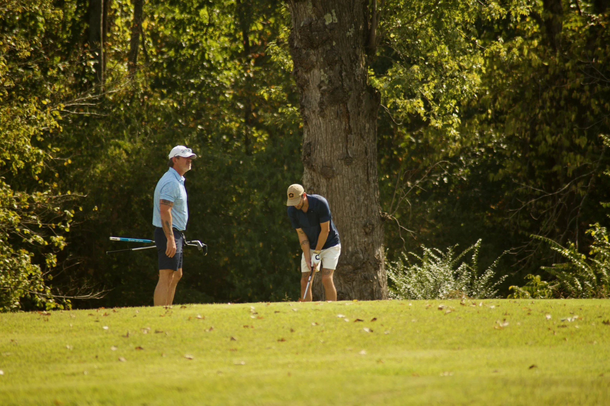 Two men playing golf on a lush green course with large trees in the background; one man is standing, watching the other who is preparing to hit a golf ball.