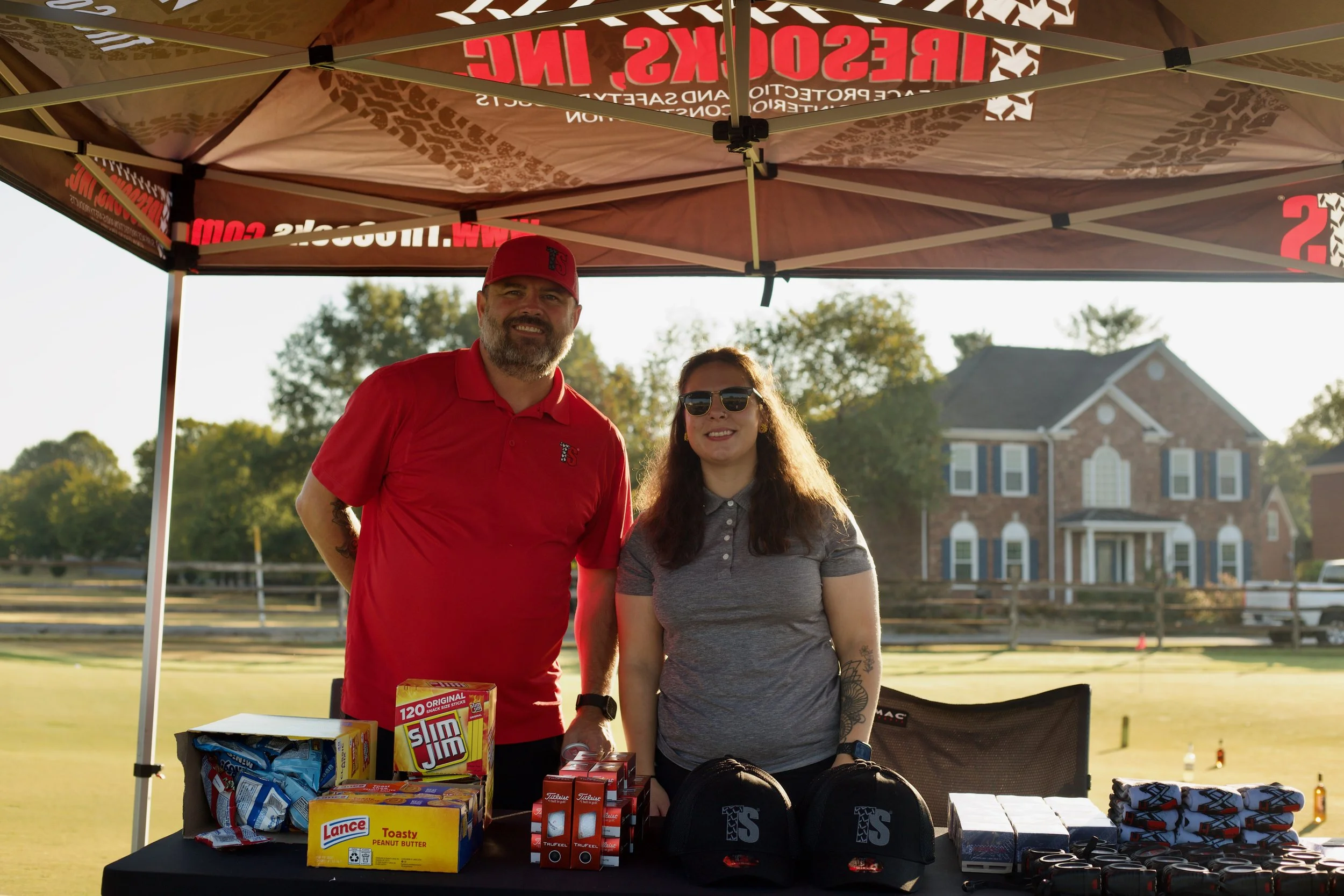 Two people standing inside a tent at an outdoor event, with tables displaying snacks and merchandise, behind a suburban house and trees during daytime.