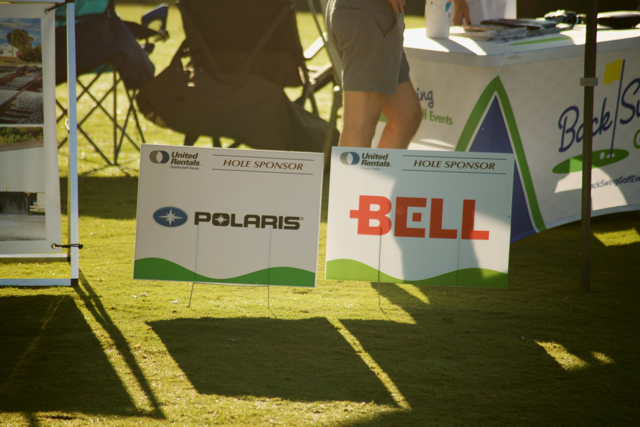 The image shows a golf event setup with signs displaying sponsor logos, including Polaris and Bell, on a grassy area with some tents or booths in the background. There are people partially visible around the booths.