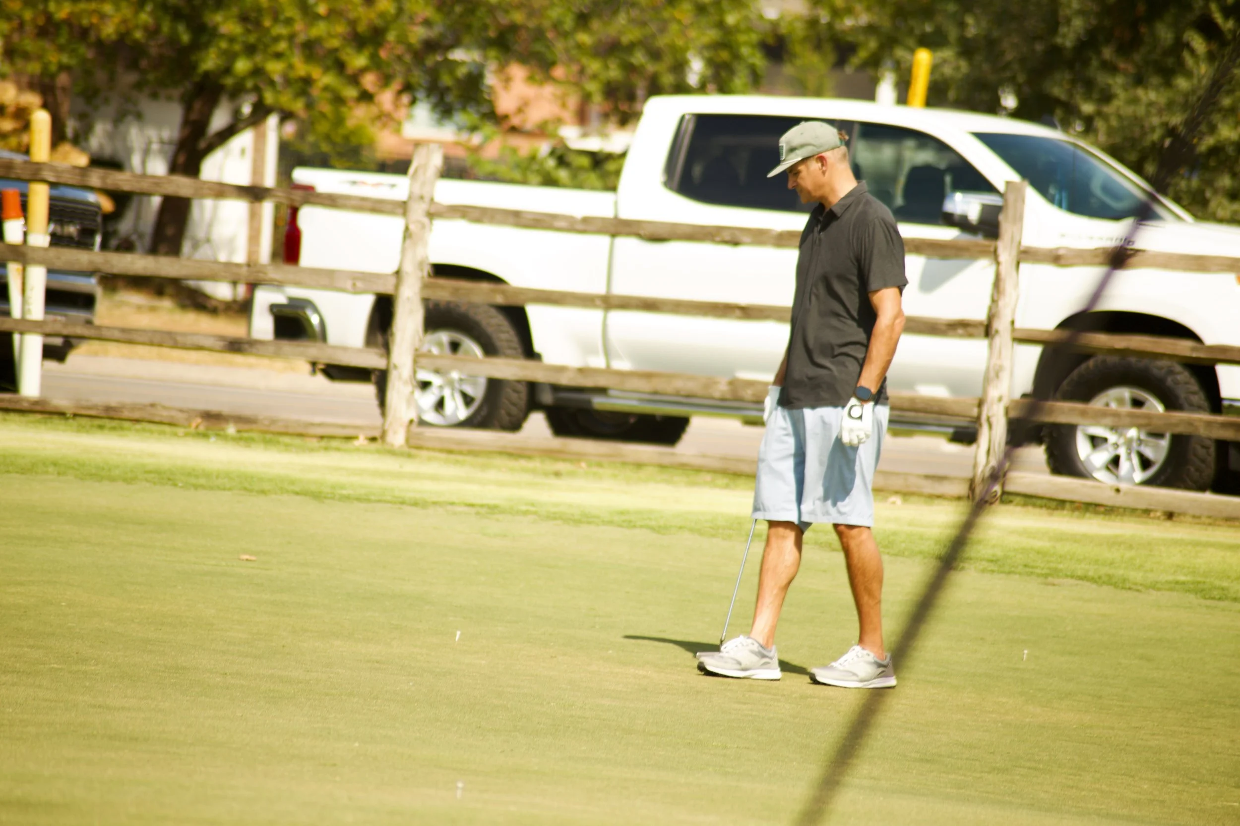 A man standing on a golf course putting, wearing a gray cap, black shirt, gray shorts, and sneakers.