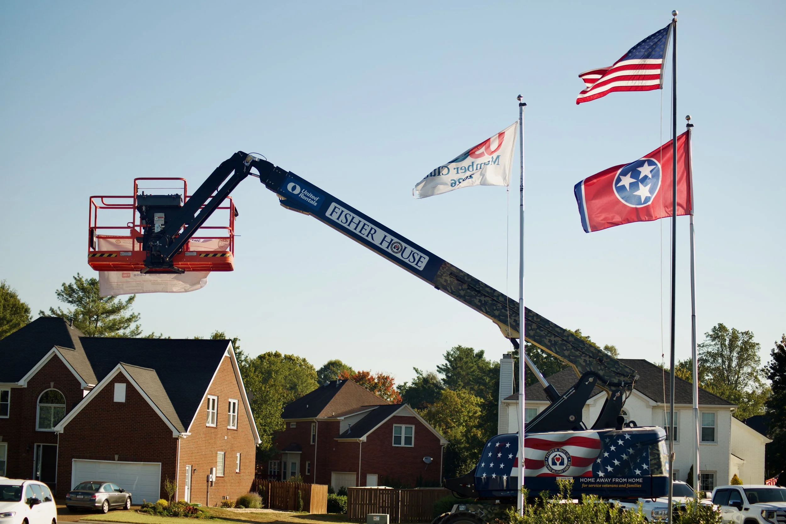 A fire truck with an extended ladder is in a residential neighborhood with brick houses and cars parked in driveways. The ladder has a platform at the top with a white bag hanging from it. The truck has American flags and a Tennessee state flag attac