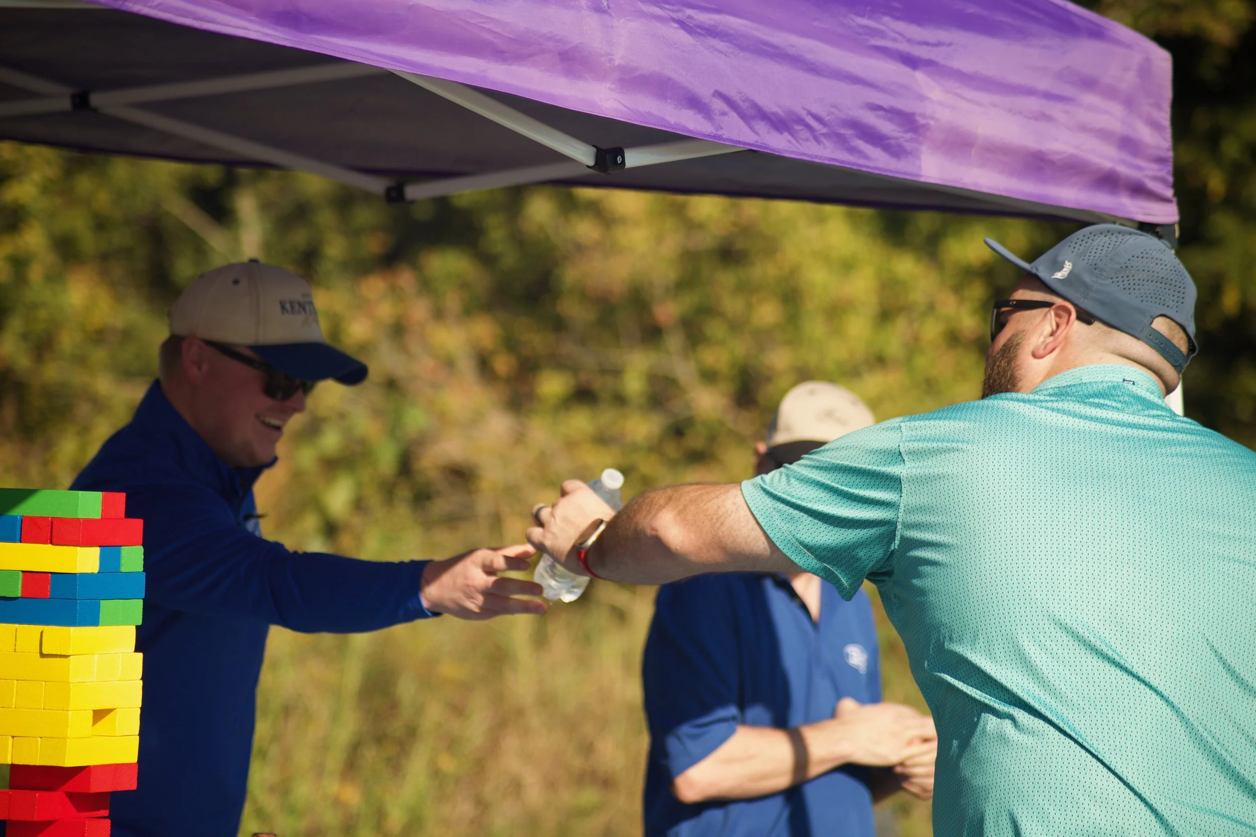 Two men shake hands at an outdoor event under a purple canopy, with a colorful Jenga tower on the left. The man on the left is wearing a beige cap and glasses, the man on the right is wearing a blue cap and sunglasses. Trees with autumn foliage are i