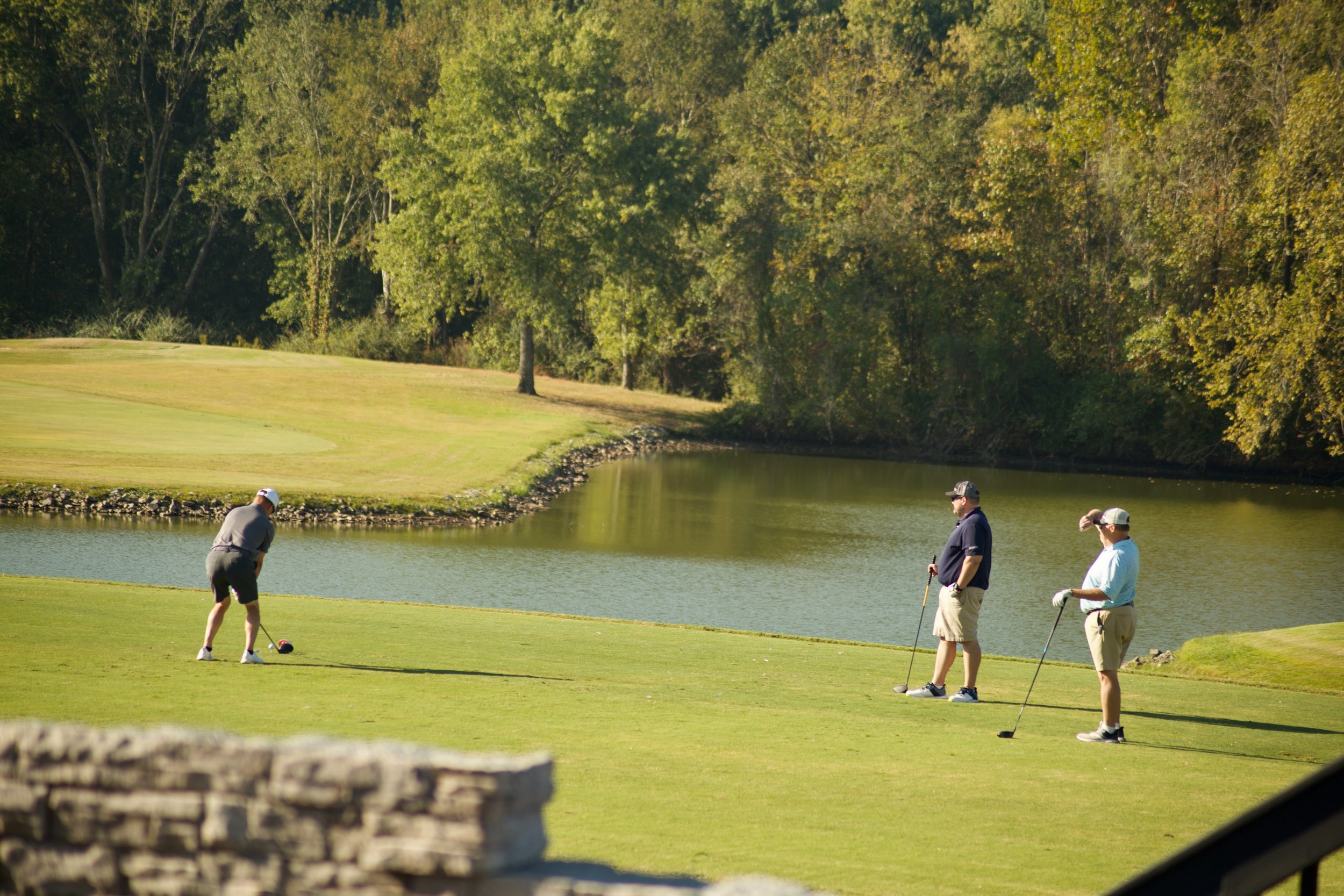 People playing golf on a course by a pond surrounded by trees.