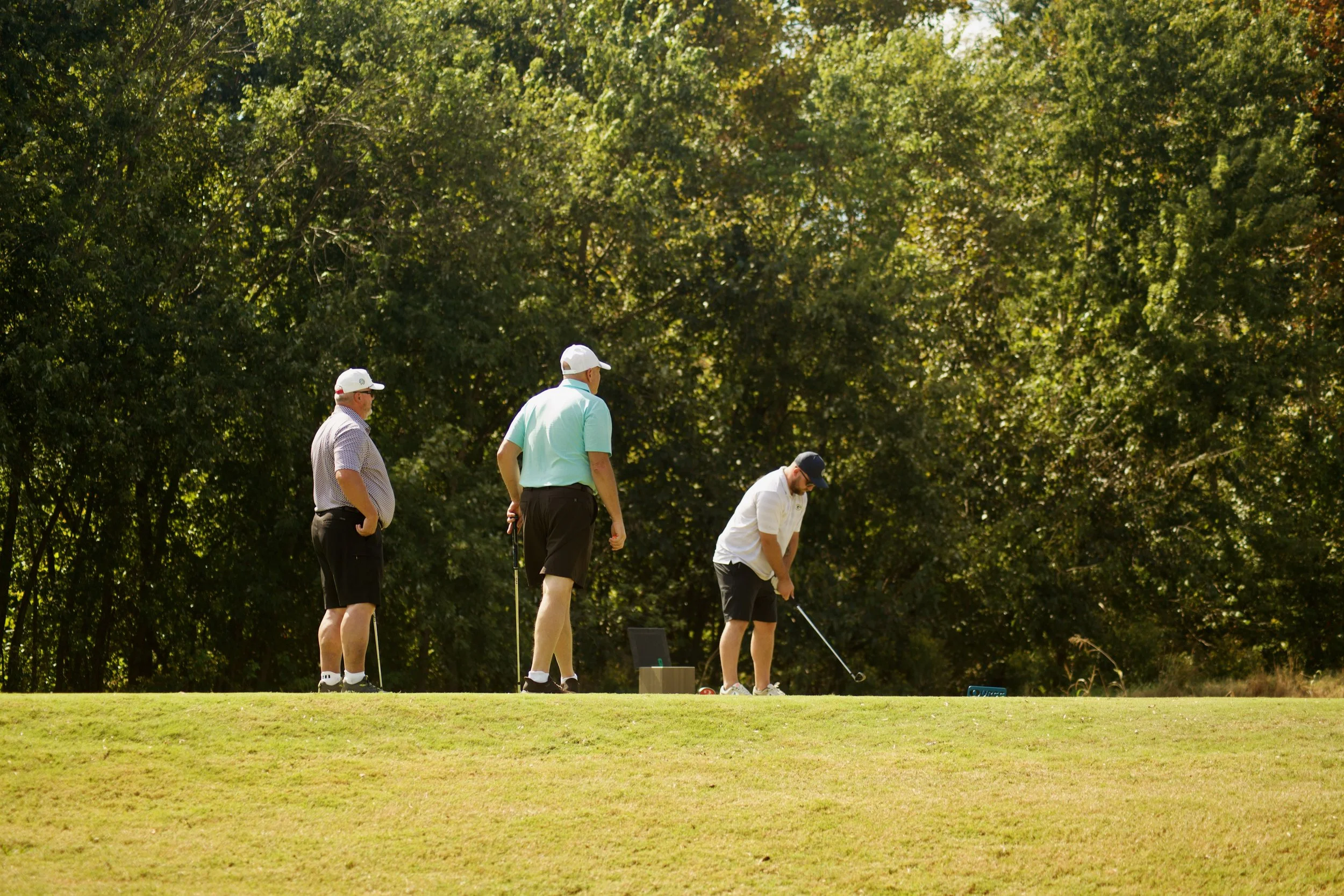 Four men playing golf on a lush green course with trees in the background, one preparing to putt, others watching.