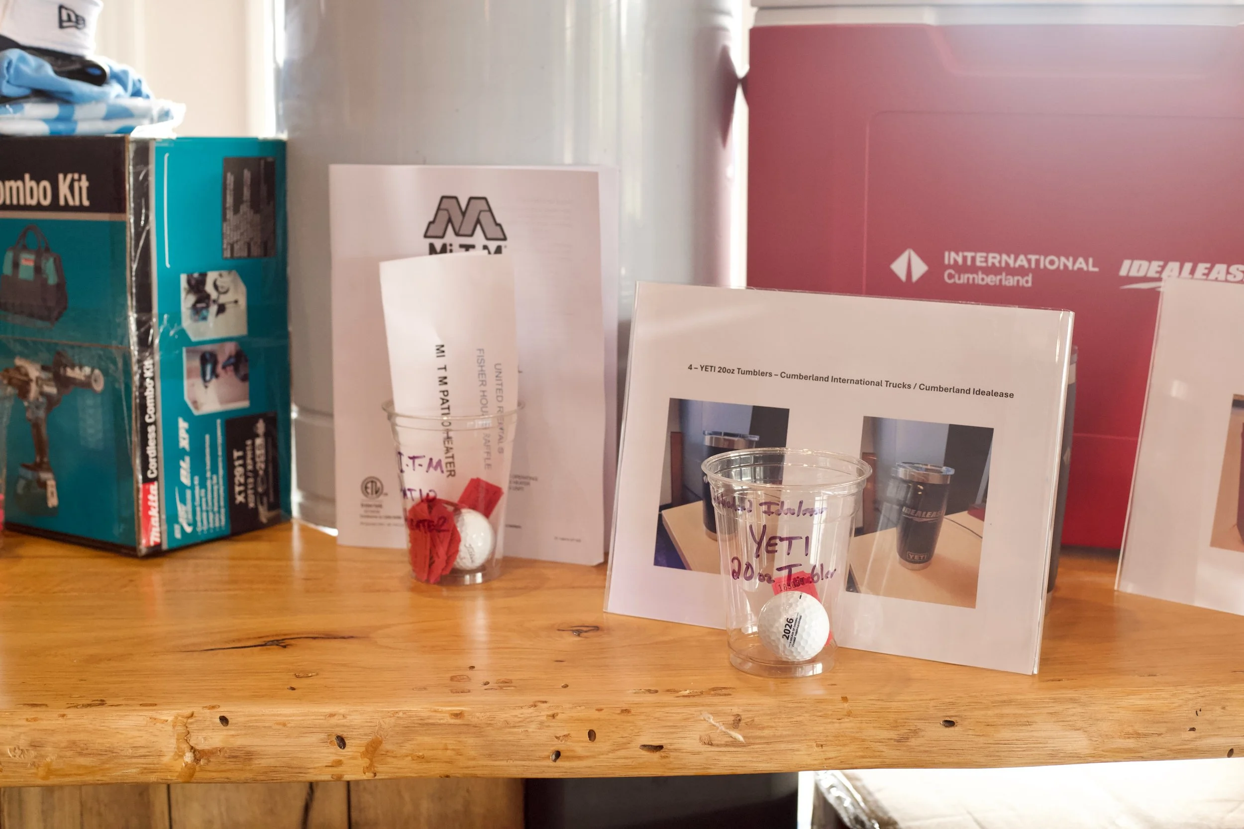 Display of donated items, including a box, brochures, and a clear plastic cup with a golf ball labeled 'YETI' inside, on a wooden table with a pink box in the background.