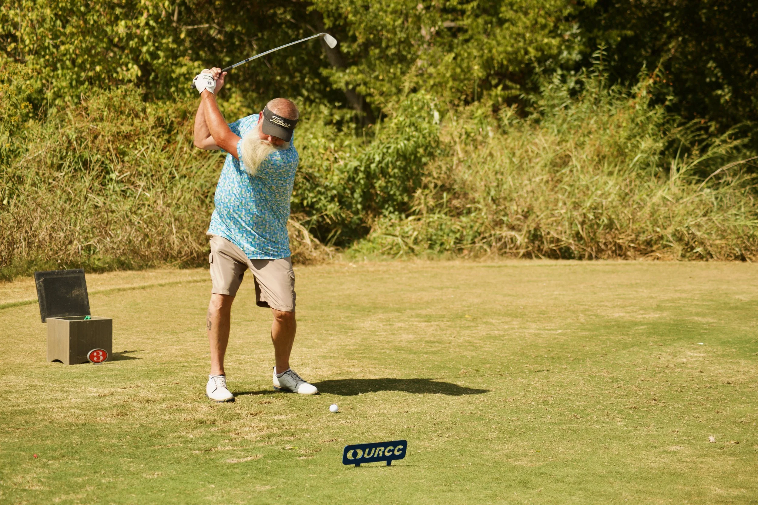 An older man with a long white beard and tattoos on his legs playing golf on a sunny day.