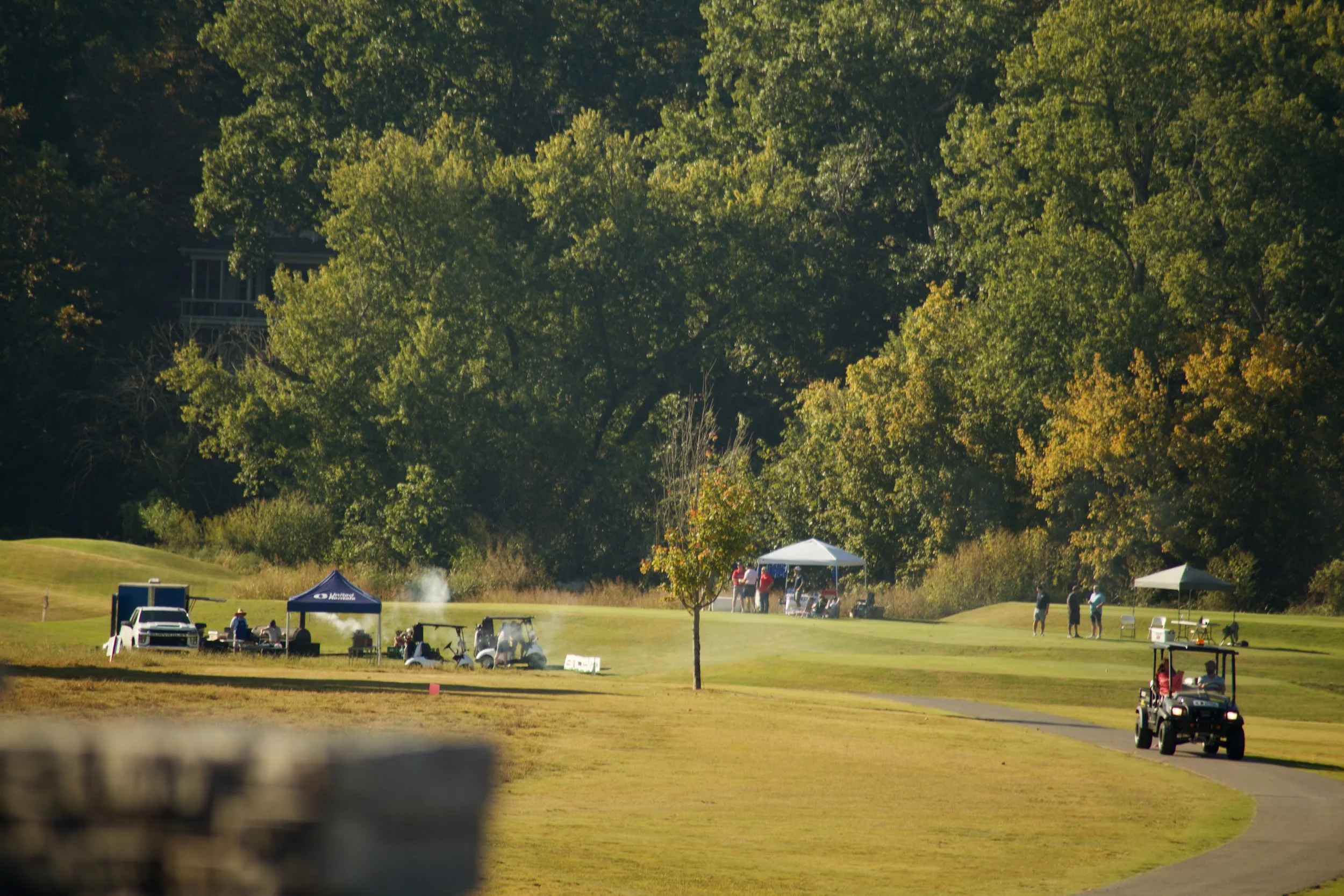 View of a golf course with people gathered under tents, surrounded by trees, with golf carts and vehicles nearby.