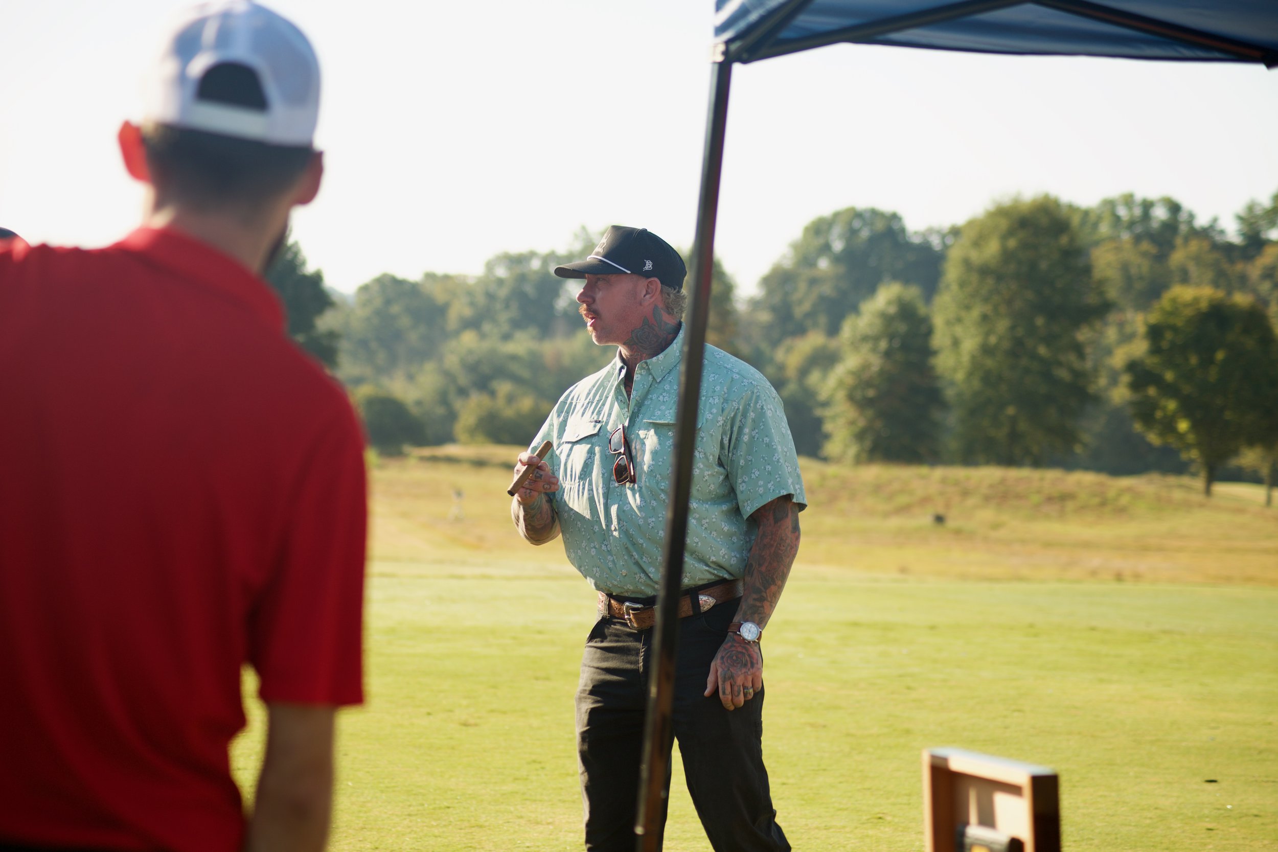 Two men talking outdoors on a sunny day, with one man in focus wearing a floral shirt, baseball cap, and tattoos, holding a cigar, while the other man is blurred in the foreground wearing a red shirt and a white cap.