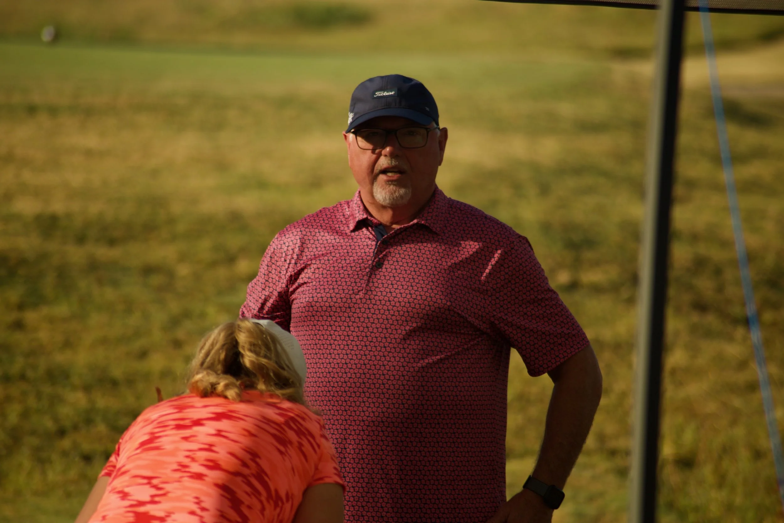 A man wearing glasses, a black cap, and a red patterned shirt standing outdoors on green grass.