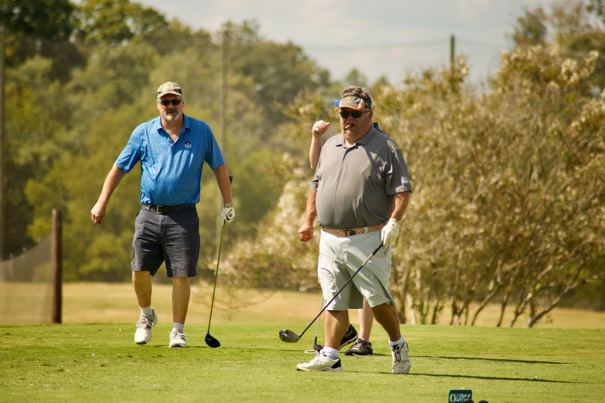 Two middle-aged men playing golf on a sunny day. One man is wearing a blue shirt, shorts, and a cap, while the other is in a grey shirt and white shorts, holding a golf club.